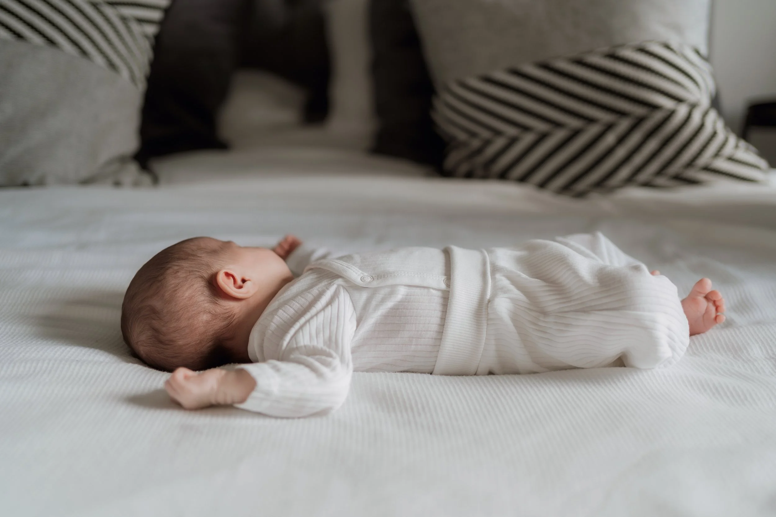 A baby lying on a white bed wearing a white outfit, with pillows in black and white stripes and solid colors in the background.