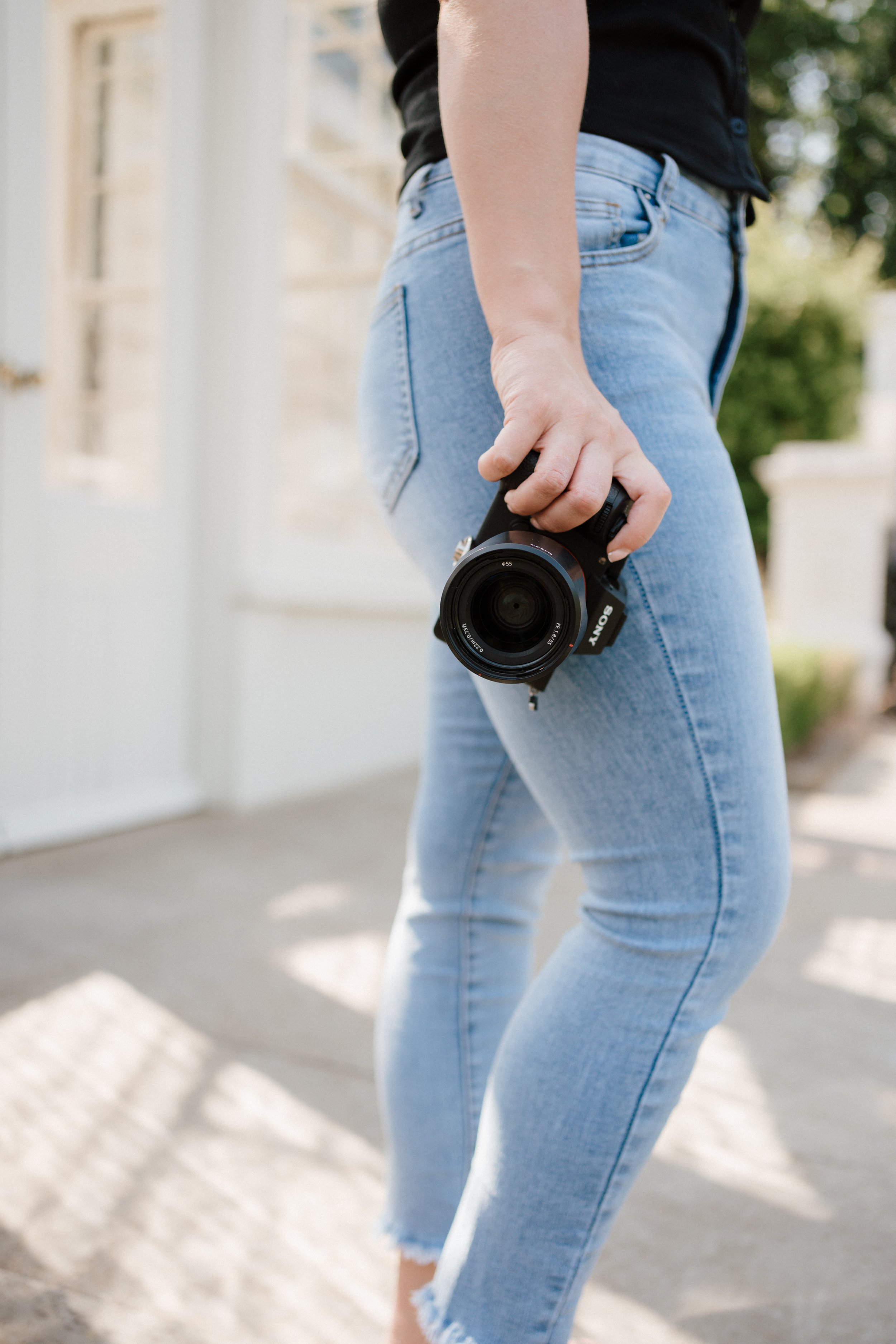 A person wearing light blue jeans and a black shirt holding a Sony camera, standing outdoors near a white building with a glass door.