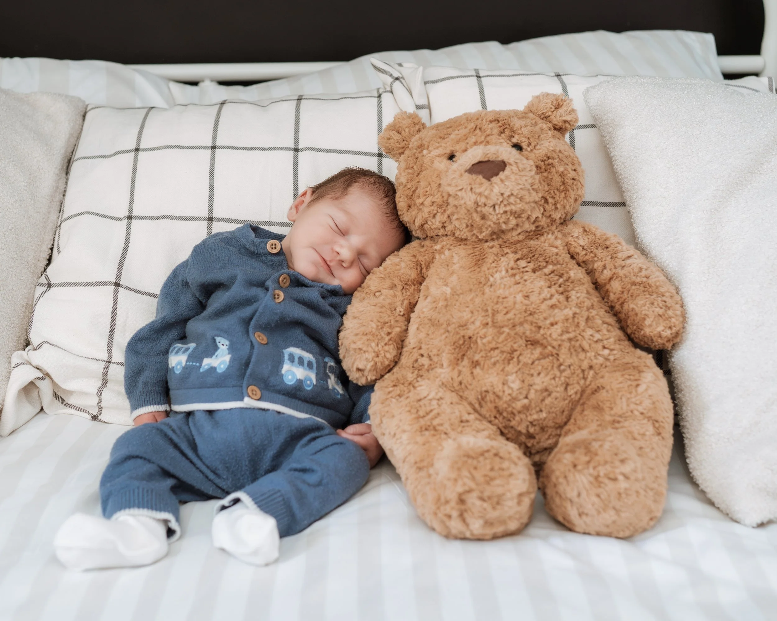 A sleeping baby laying on a bed next to a large teddy bear with pillows around them.