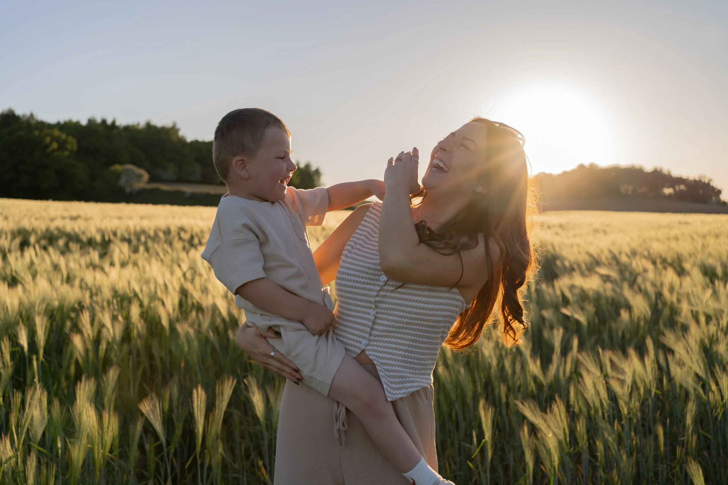 A woman and a young boy enjoying a playful moment in a wheat field during sunset, with the woman holding the boy in her arms and both laughing.