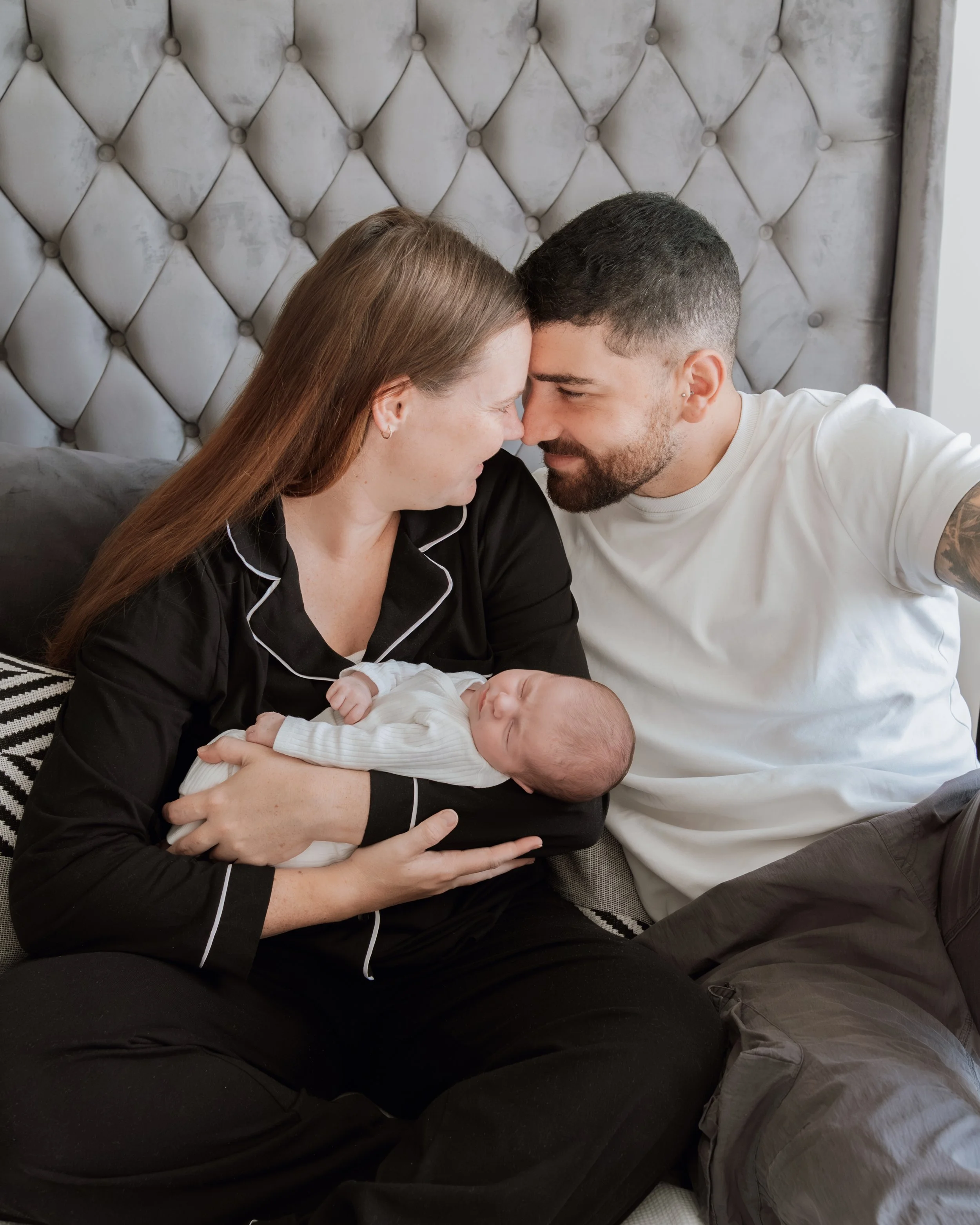 A woman is holding a sleeping newborn baby while a man and woman lean in towards each other, touching foreheads, in a cozy bedroom with a tufted headboard.
