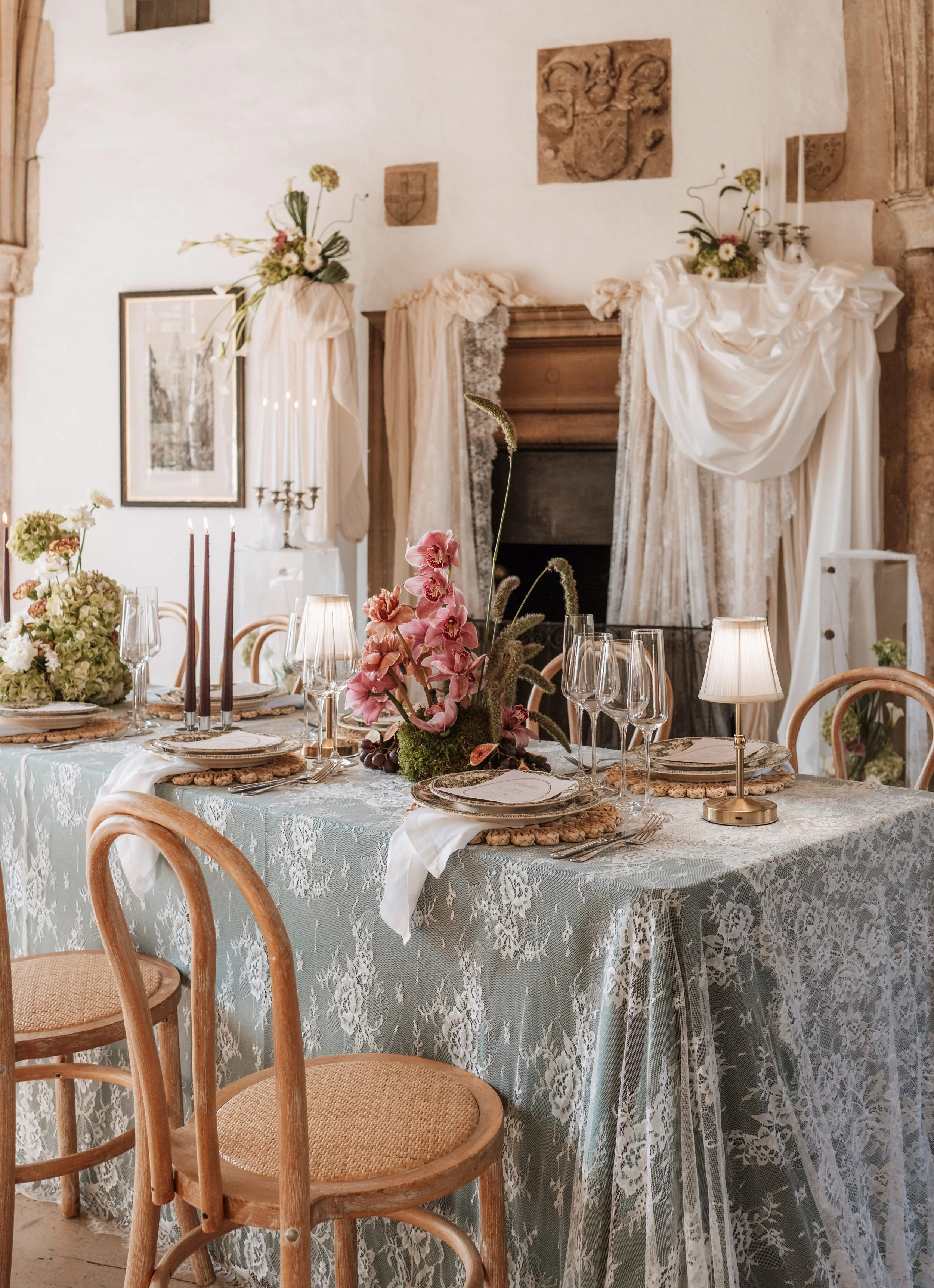 Elegant dining table decorated with floral centerpieces, tableware, candles, and small lamps in a rustic elegant setting with draped fabric and wall decor.