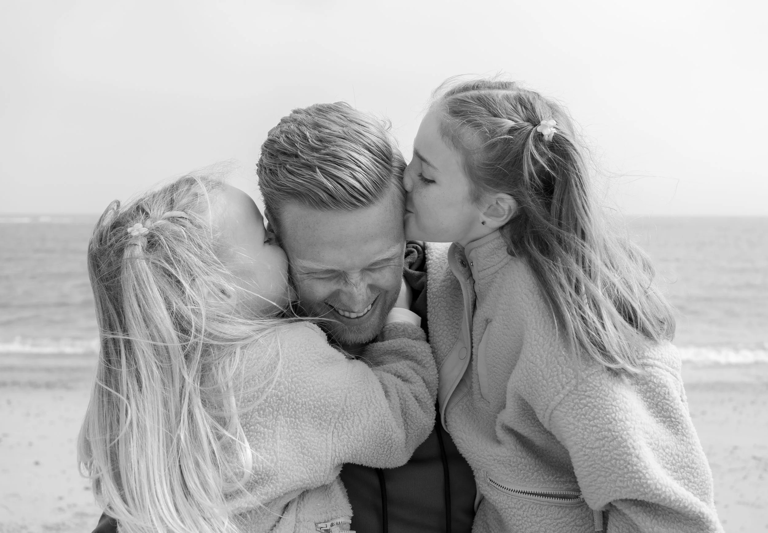 A man with two young girls, one on each side, in an affectionate moment at the beach. The girls kiss his cheeks as he smiles with eyes closed.