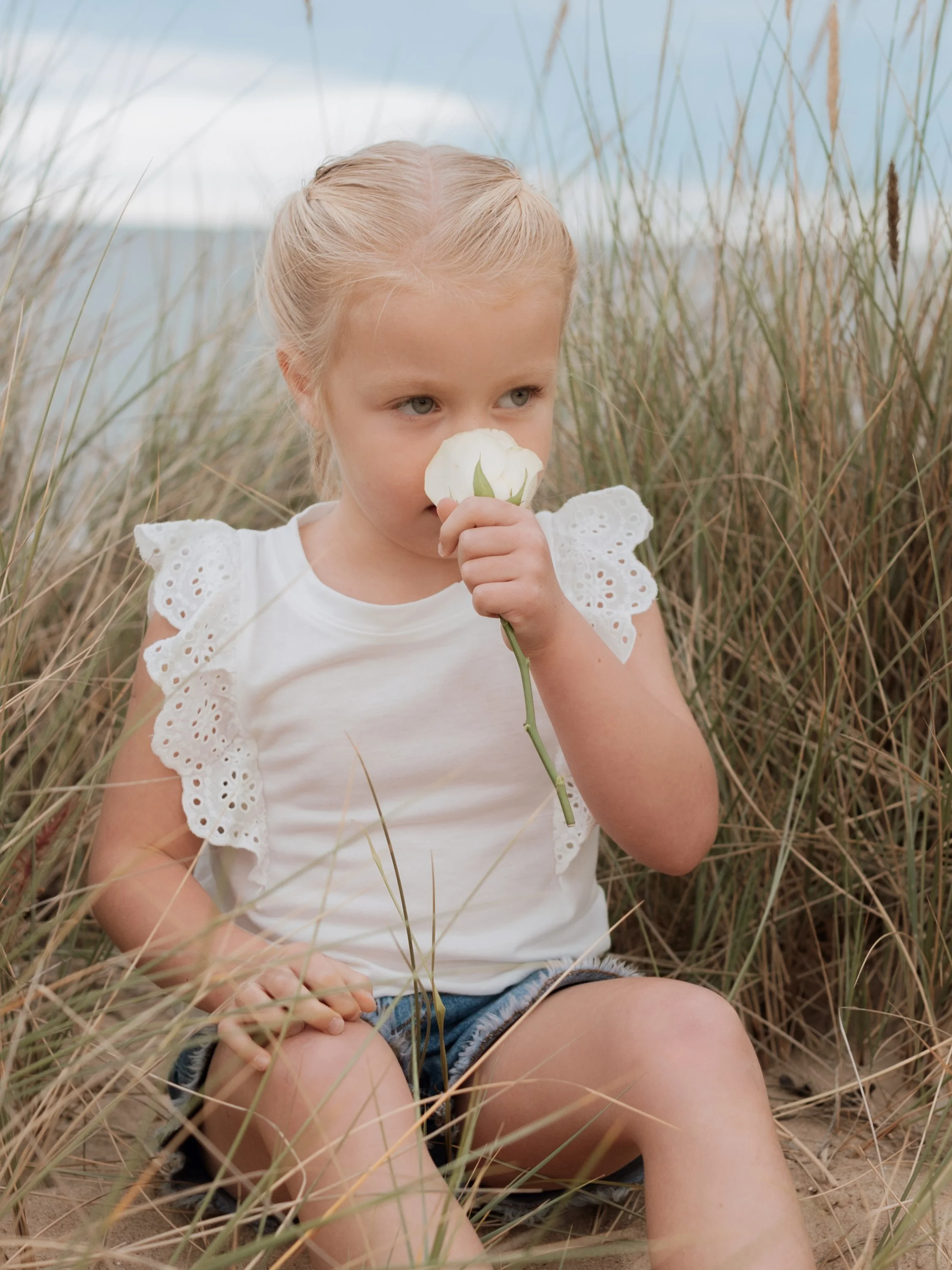A young girl with blonde hair, wearing a white top with lace sleeves and denim shorts, sitting in tall grass and holding a white flower close to her nose.