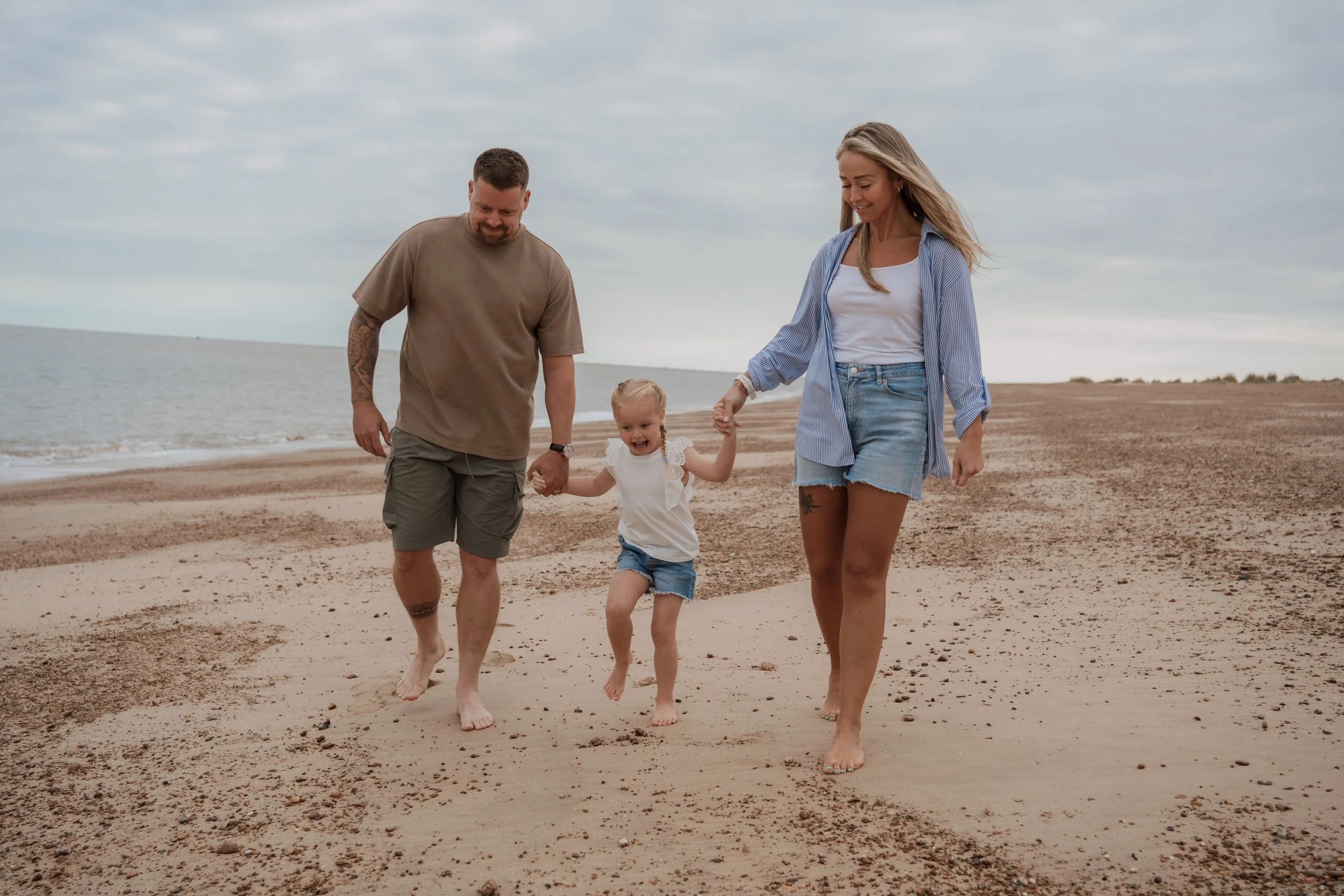 A family of three walking barefoot on the beach, holding hands, with the ocean and cloudy sky in the background.