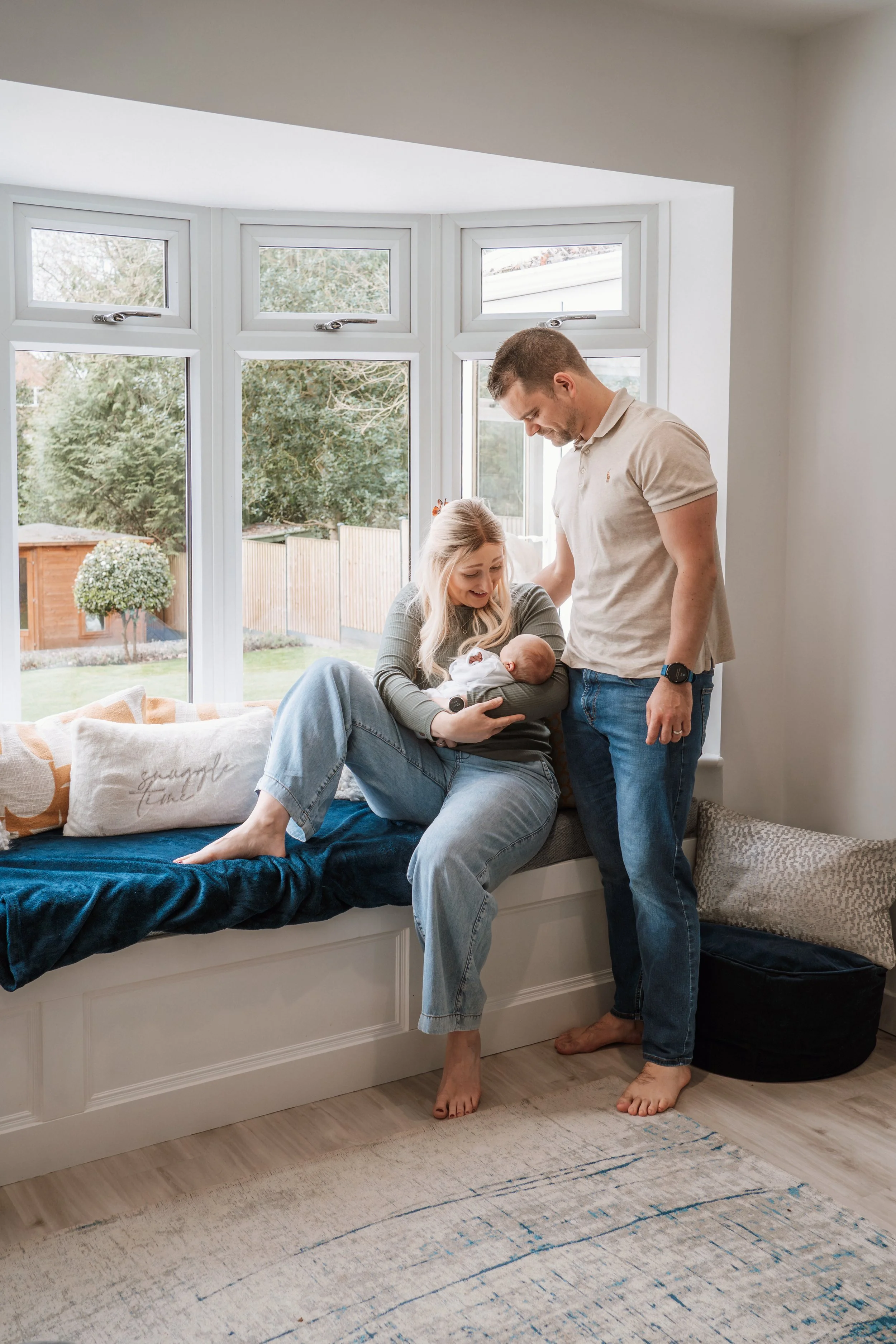 A woman holding a newborn baby, with a man standing next to her, in a bright sitting area by large bay windows, with a garden visible outside.