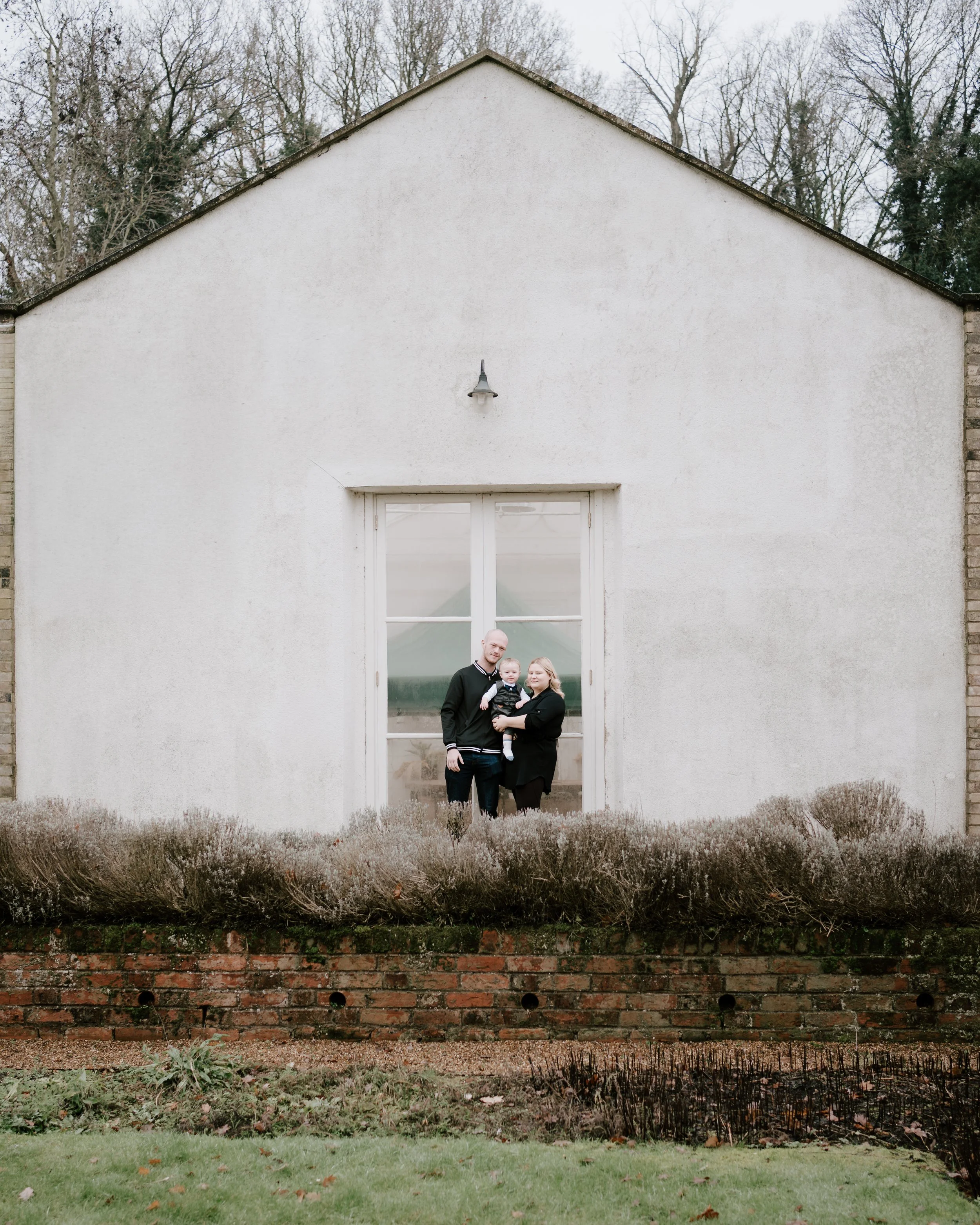 A family of three standing in front of a large window on a white house, with trees in the background and bushes in the foreground.