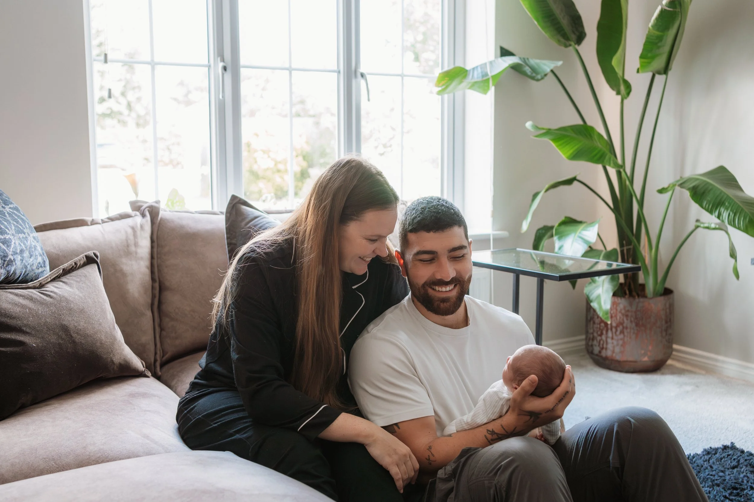 Family sitting on a sofa in a bright living room, holding a newborn, smiling and enjoying a happy moment.
