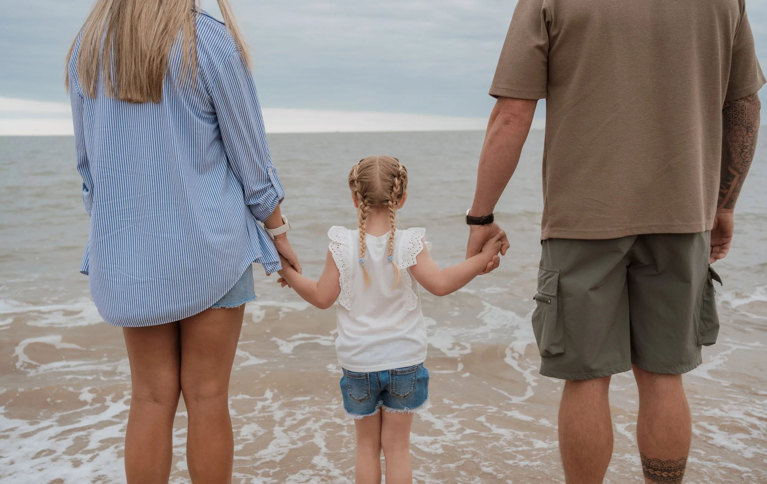 A young girl holding hands with two adults, standing on the beach facing the water.
