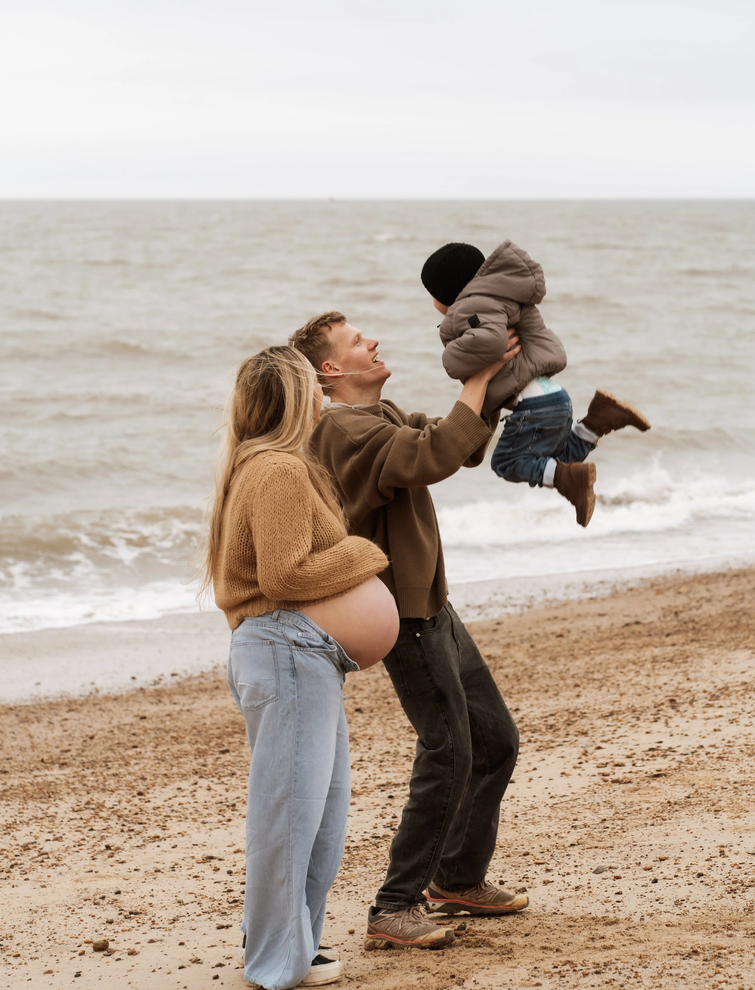 A couple on the beach holding their child in the air, with one woman showing a pregnant belly.