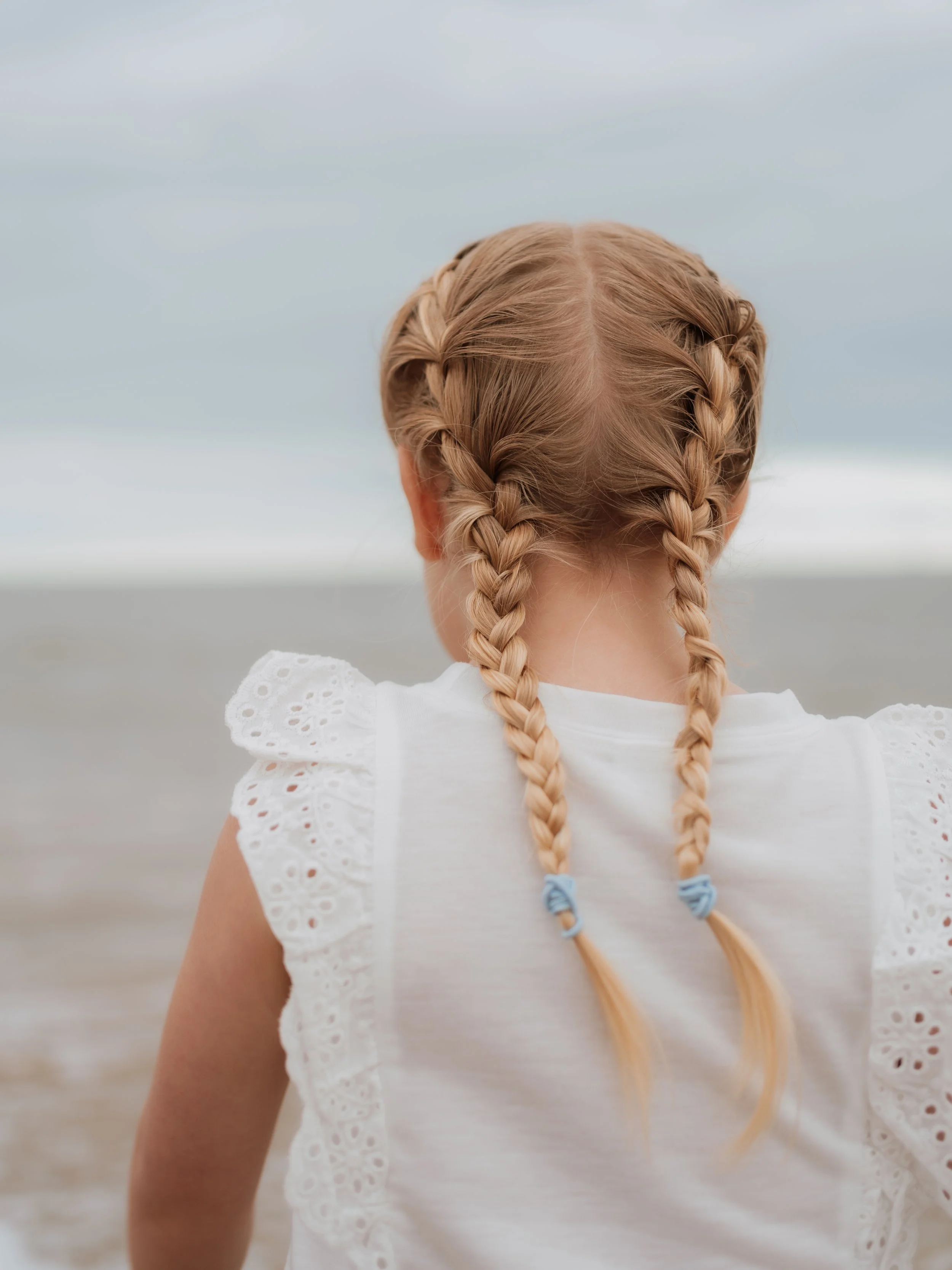 A young girl with blonde hair styled in two braids tied with light blue hair ties standing on a beach, facing towards the ocean and cloudy sky.