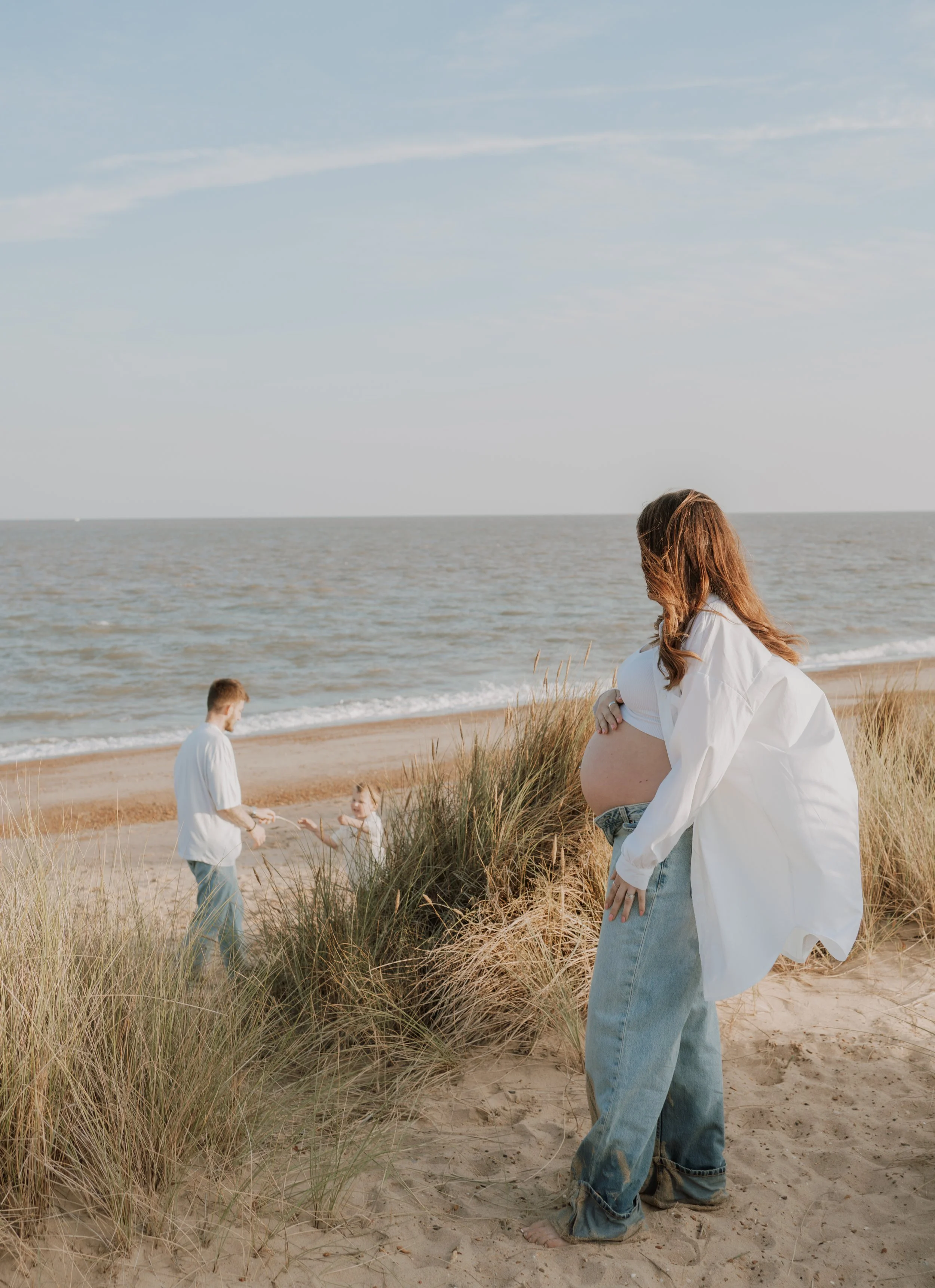 A pregnant woman on a beach with her family, including a man and a young girl playing near sand dunes and tall grass, with the ocean in the background.