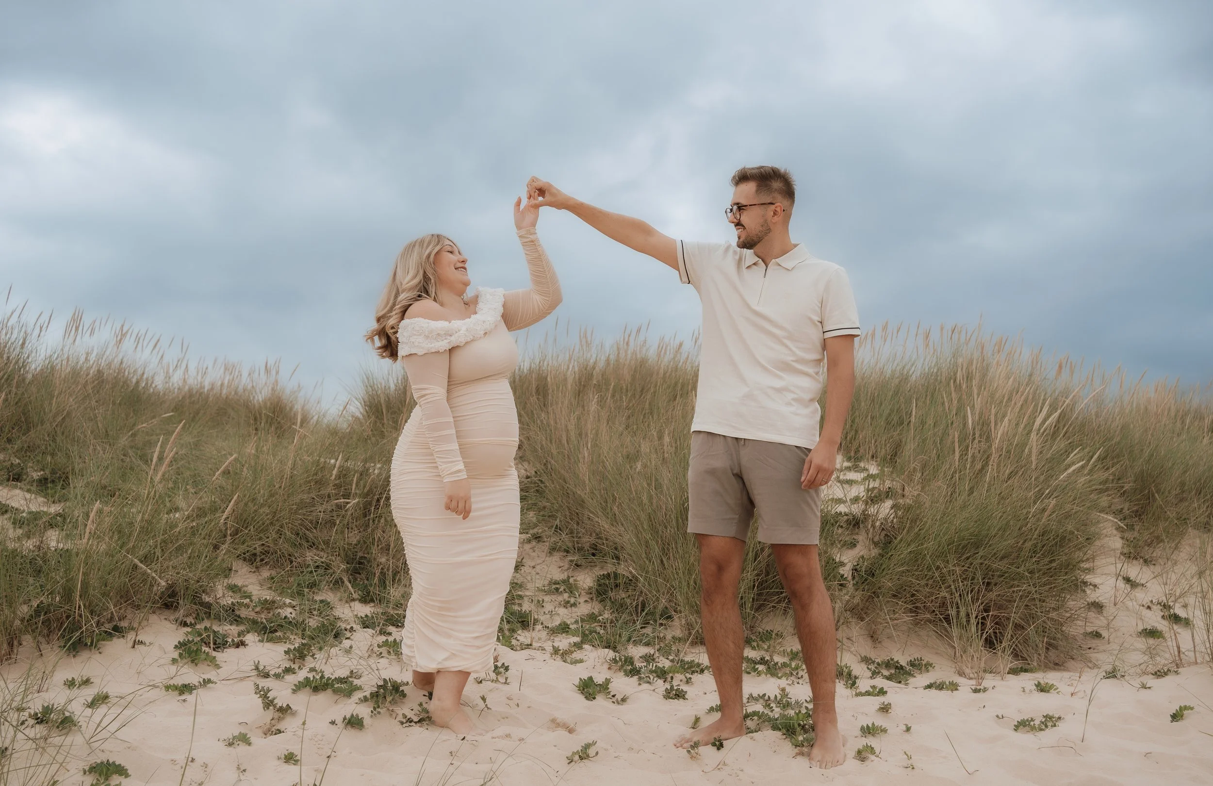 A couple dancing on a sandy beach with grass dunes and a cloudy sky in the background.