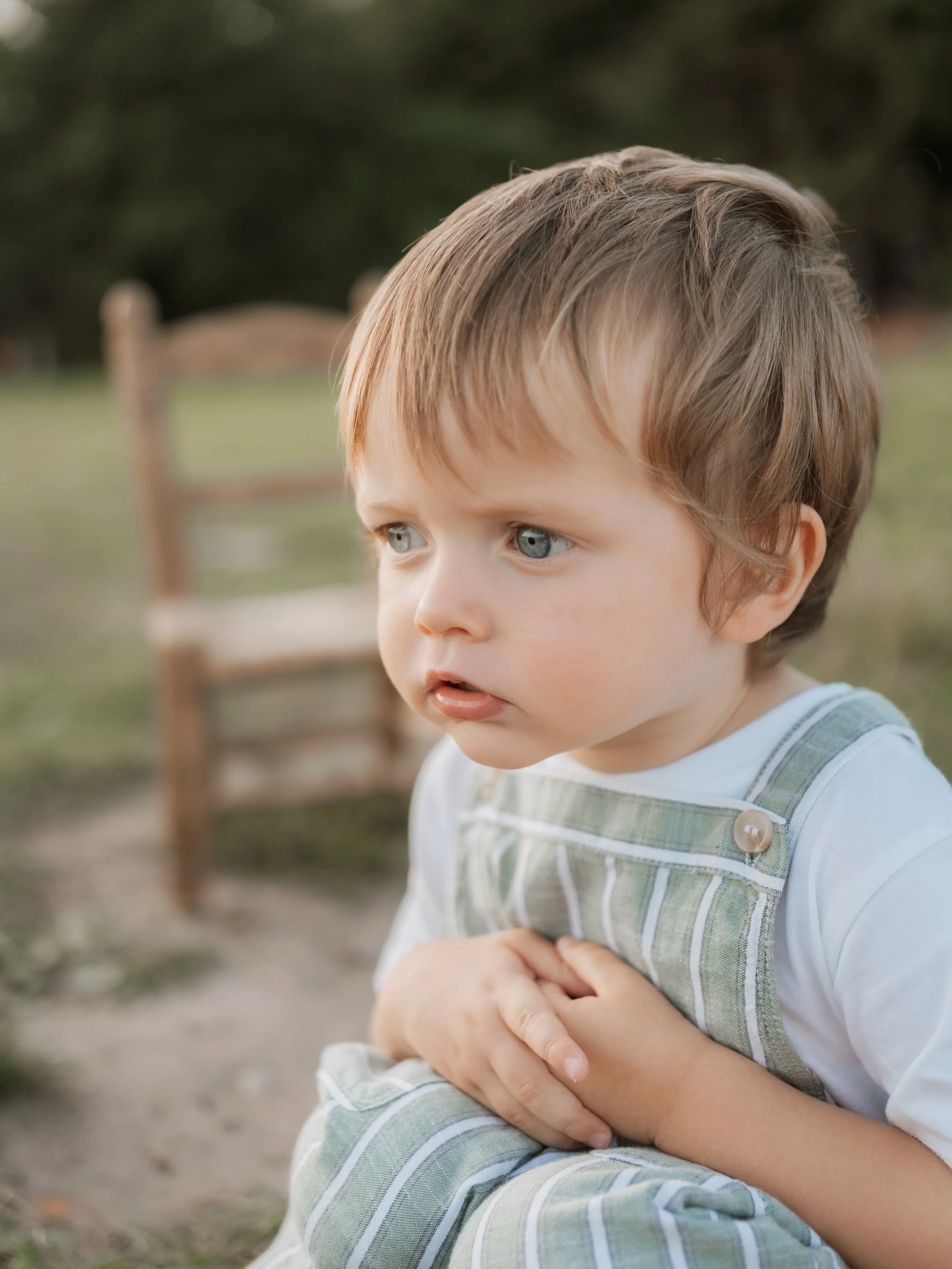 Close-up of a young boy with blue eyes and light brown hair, wearing a white shirt and green striped overalls, sitting outdoors with greenery and a wooden bench in the background.