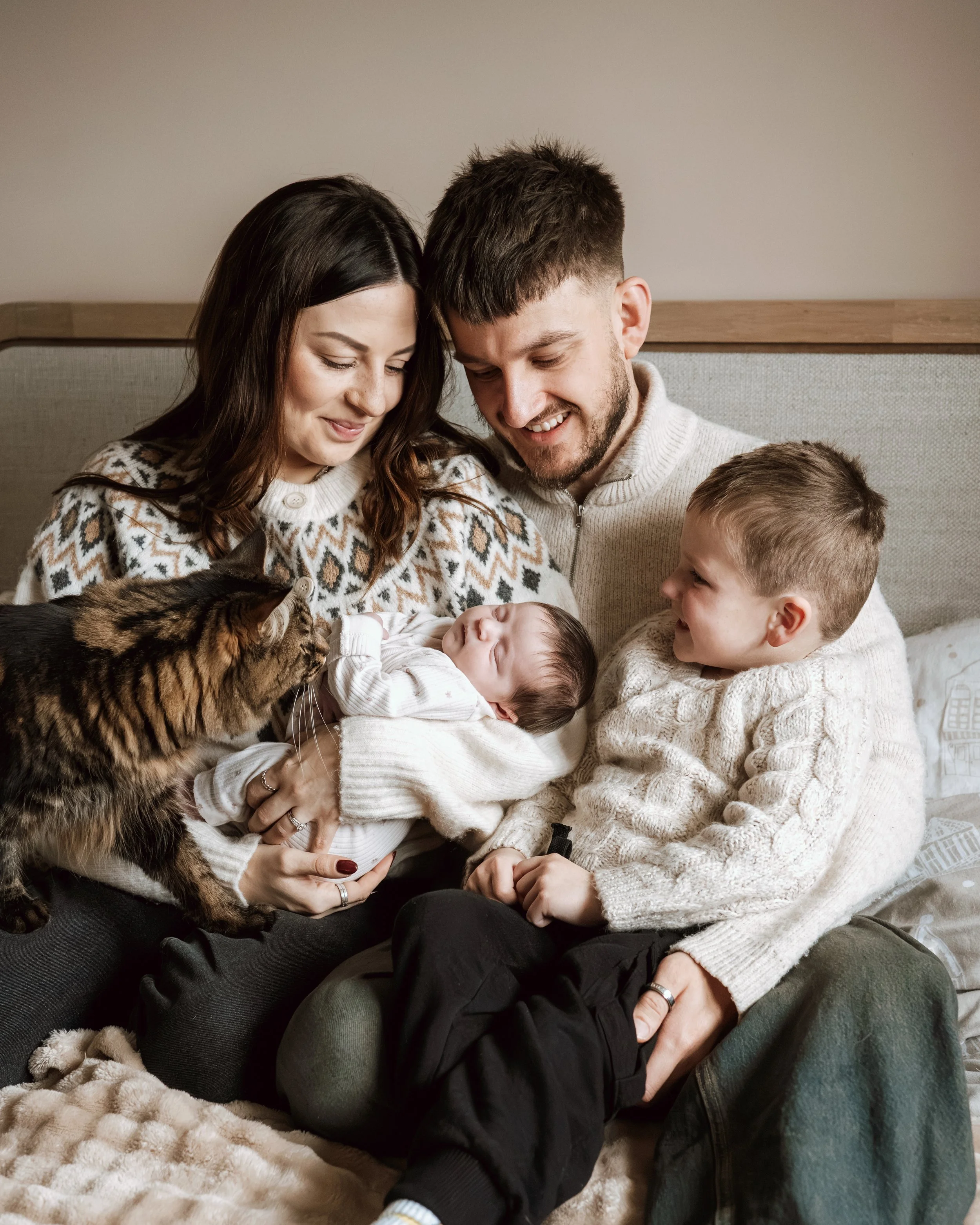 A family of four, including a mother, father, young boy, and newborn baby, sitting together on a bed, with a cat nearby, in a cozy indoor setting.