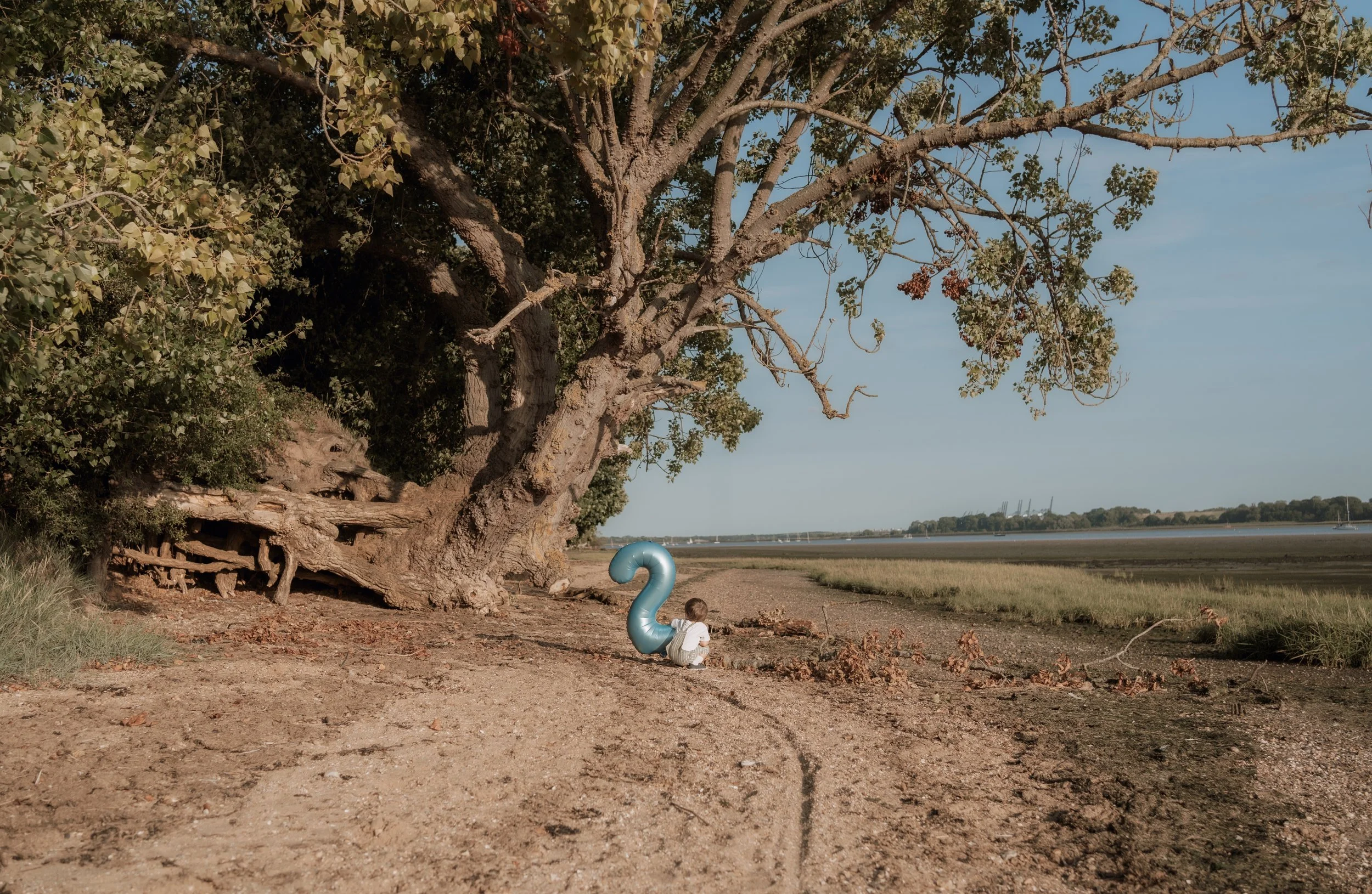 A child sitting on the ground near a large tree, holding a blue balloon shaped like the number two, on a dirt path along a coast with water and grassy land in the background under a blue sky.
