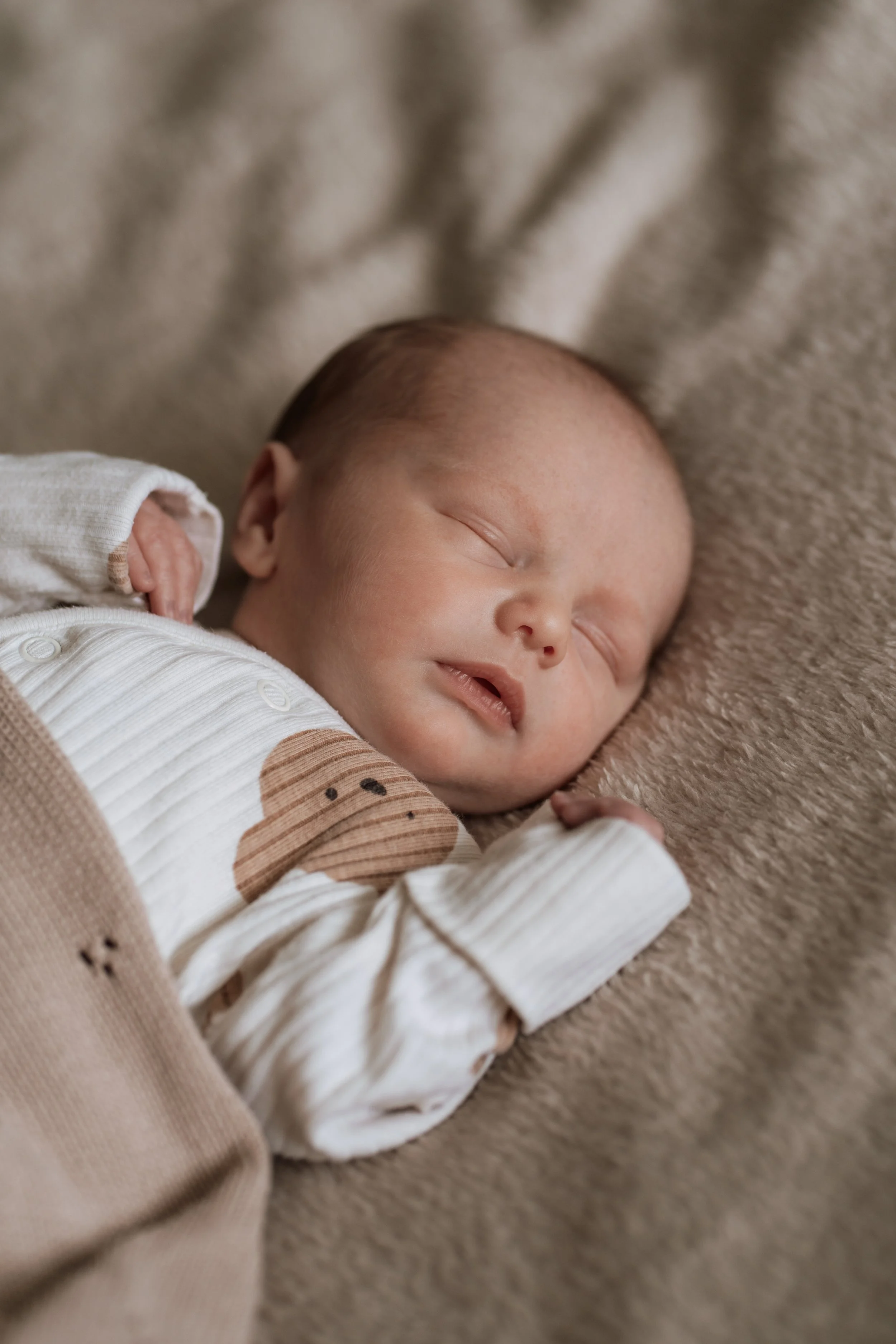 A sleeping baby with light skin and short hair, lying on a soft beige blanket, wearing a cream and brown outfit with a bear face design.