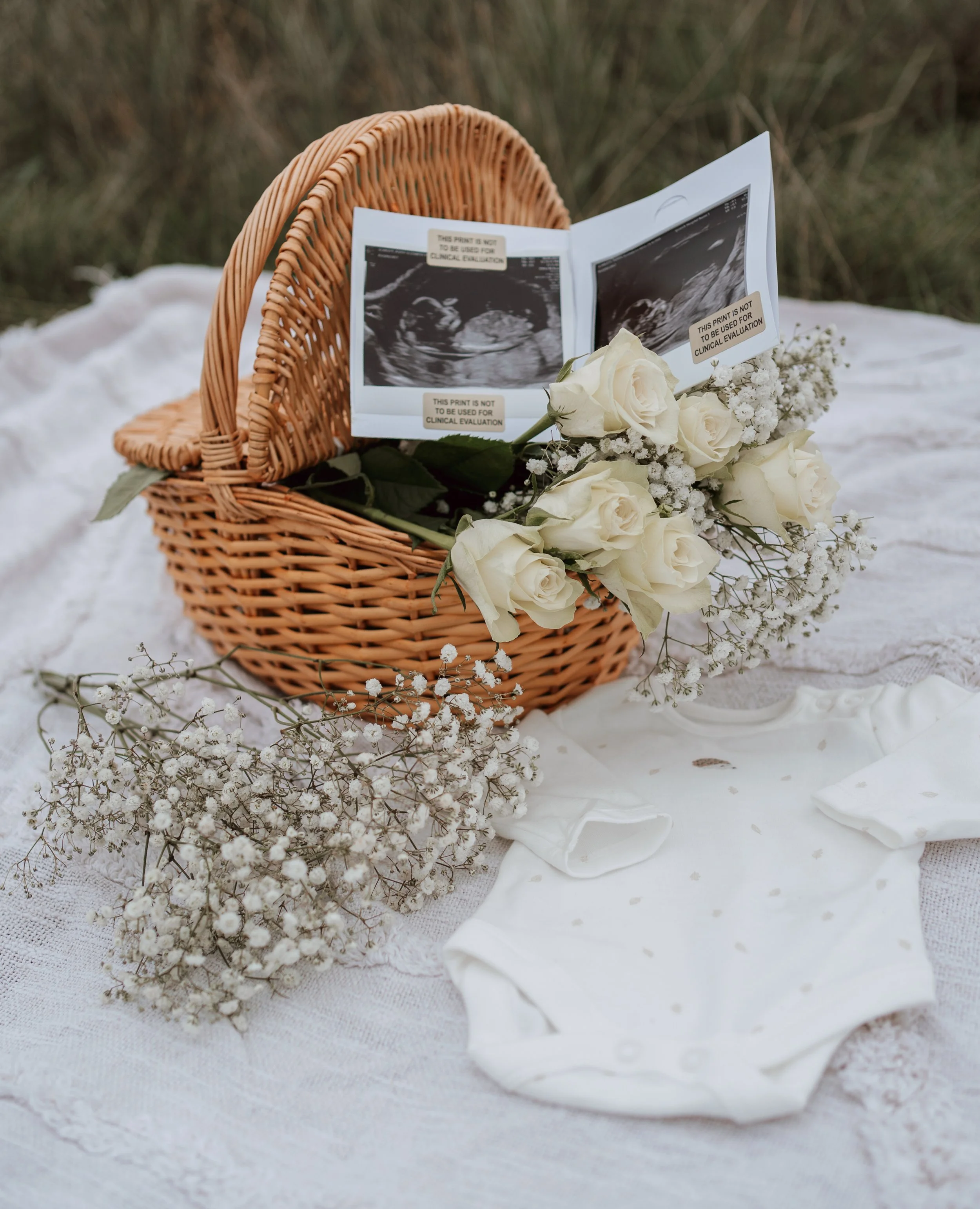 Baby ultrasound images, white roses, baby's onesie, and baby’s breath flowers arranged on a white cloth, in a wicker basket.