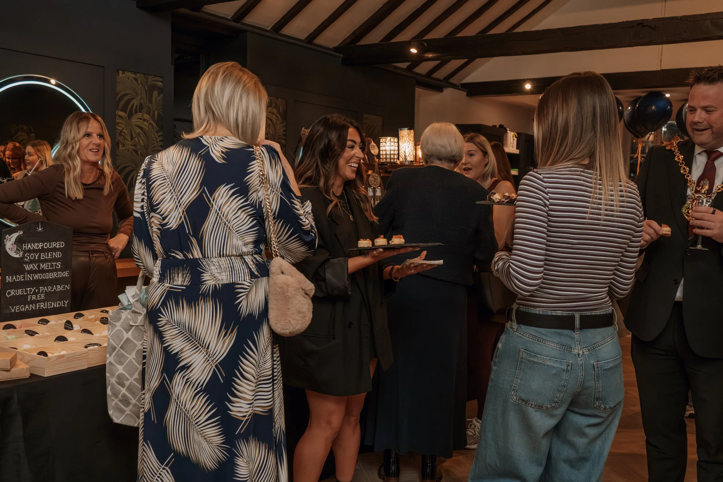 Group of people socializing at an indoor event, with some holding trays of small food items, in a warmly lit room with dark walls and ceiling beams.