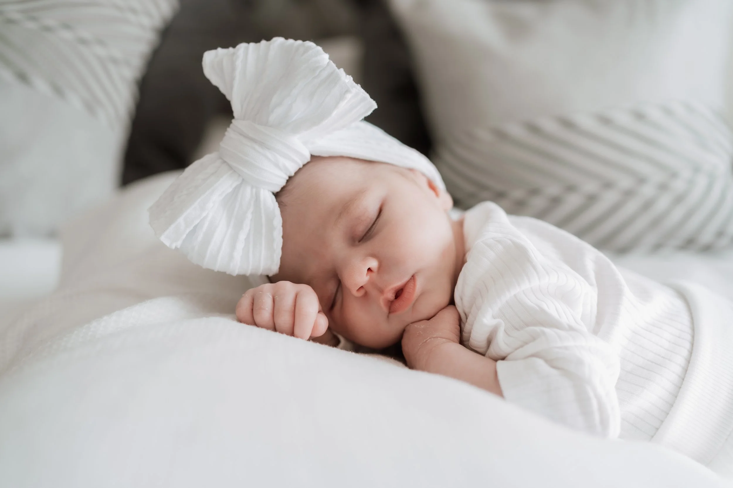 A sleeping baby with a white bow headband, resting on a soft surface, surrounded by pillows.