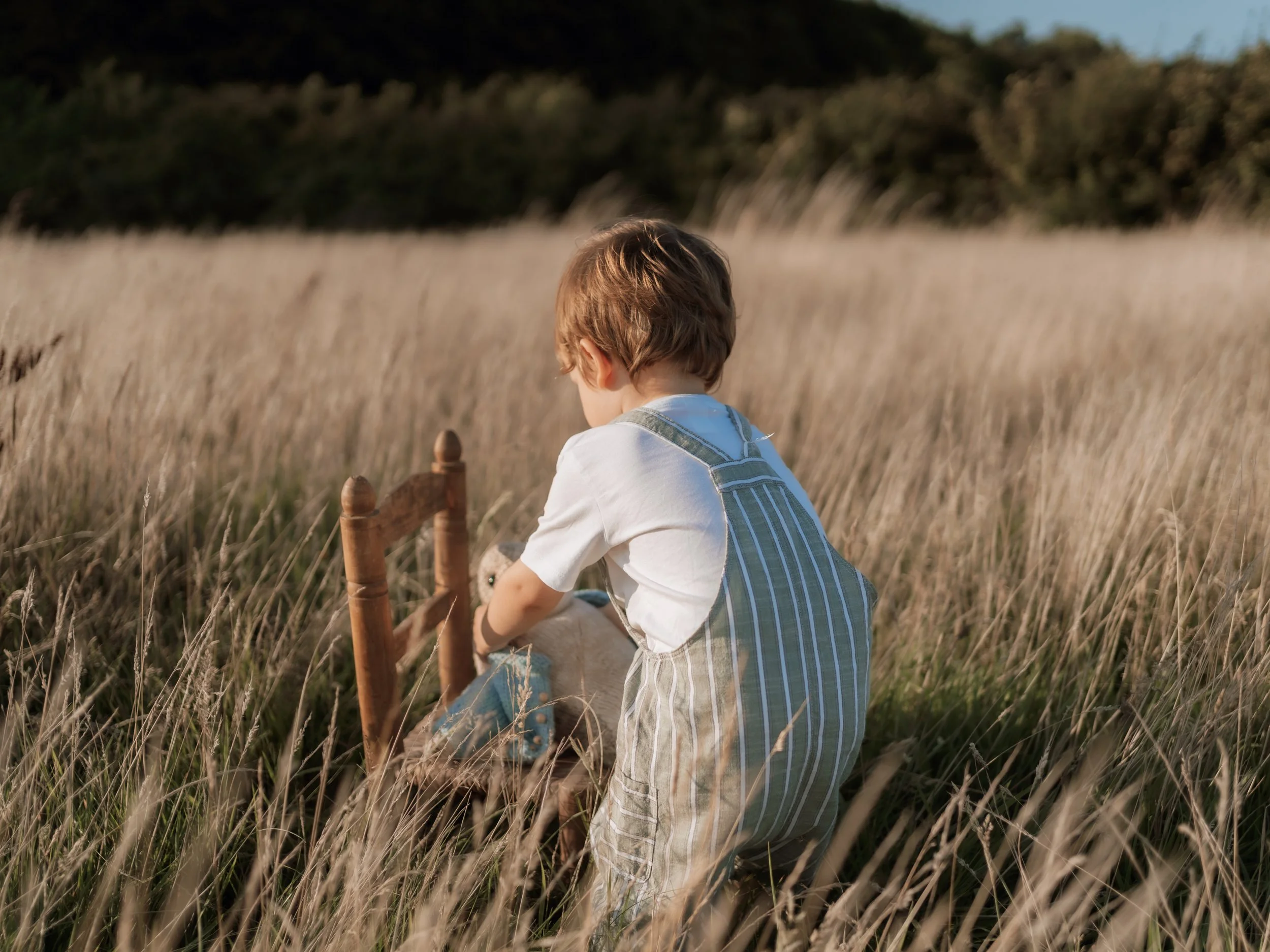 A young boy in striped overalls and a white shirt playing with a small wooden chair in a field of tall, dry grass.