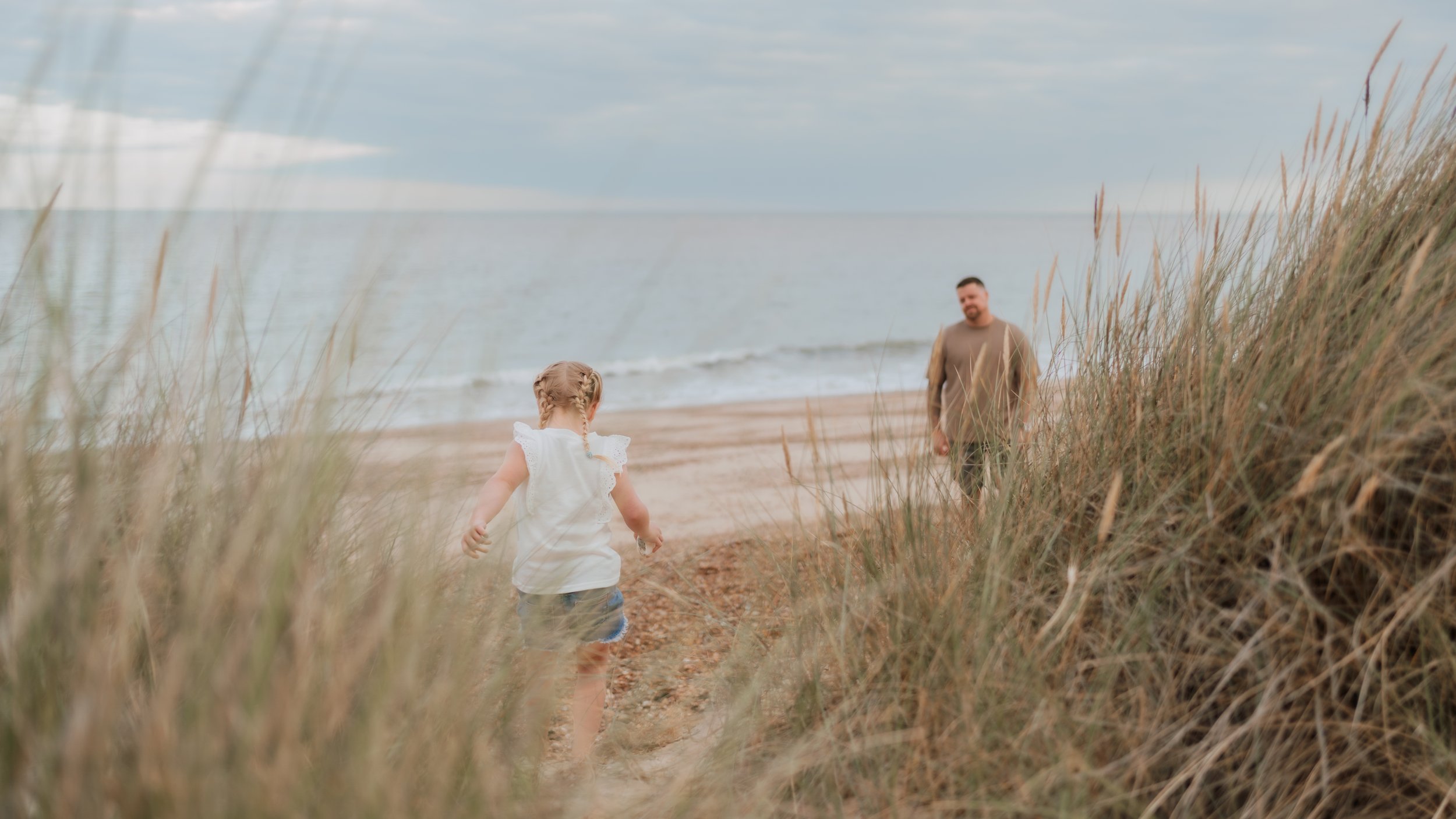 A young girl with braided hair walking through tall grass on a beach toward a man by the water in the distance.