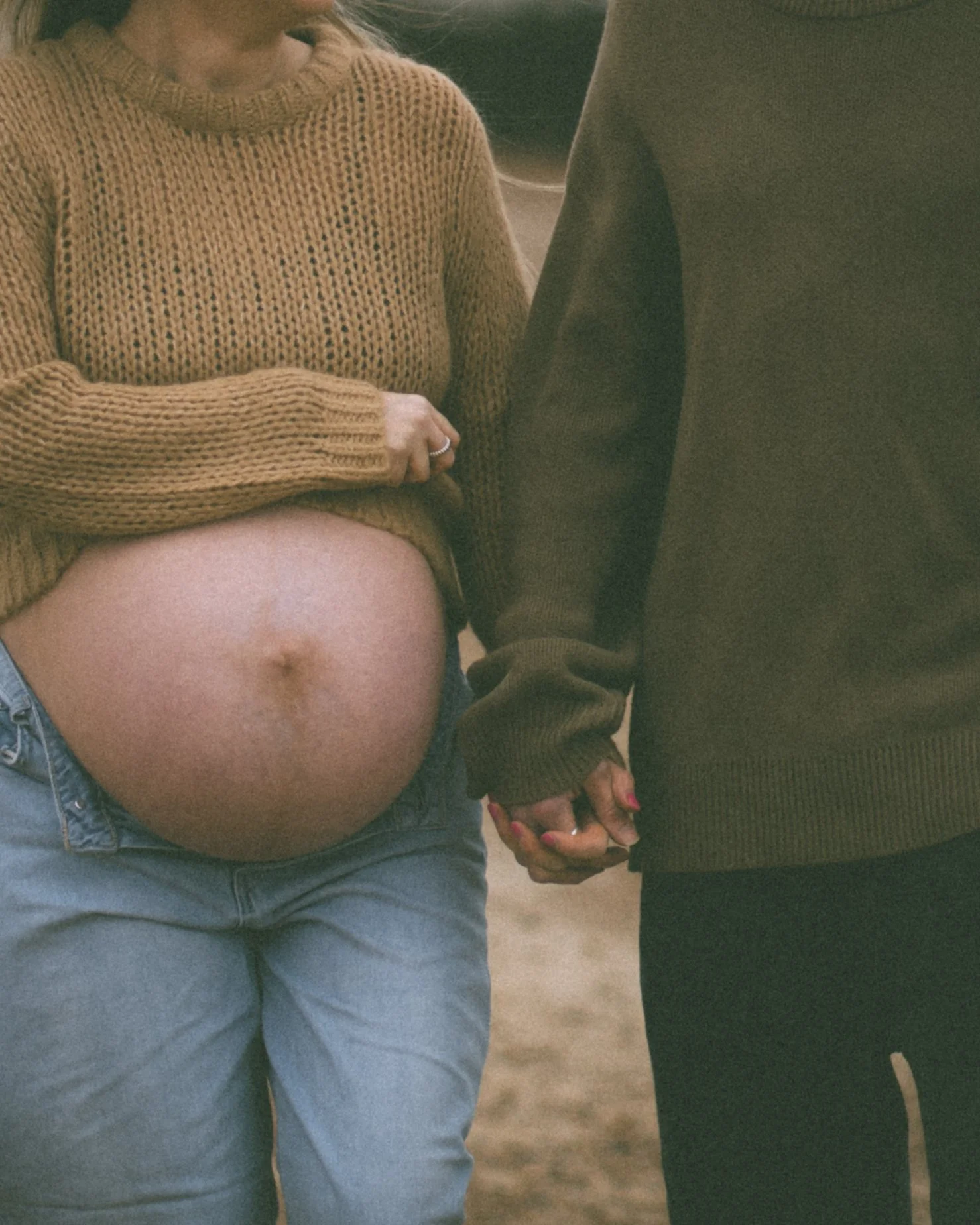 A pregnant woman holding hands with her partner, showing her bare belly, wearing a mustard sweater and jeans, outdoors.