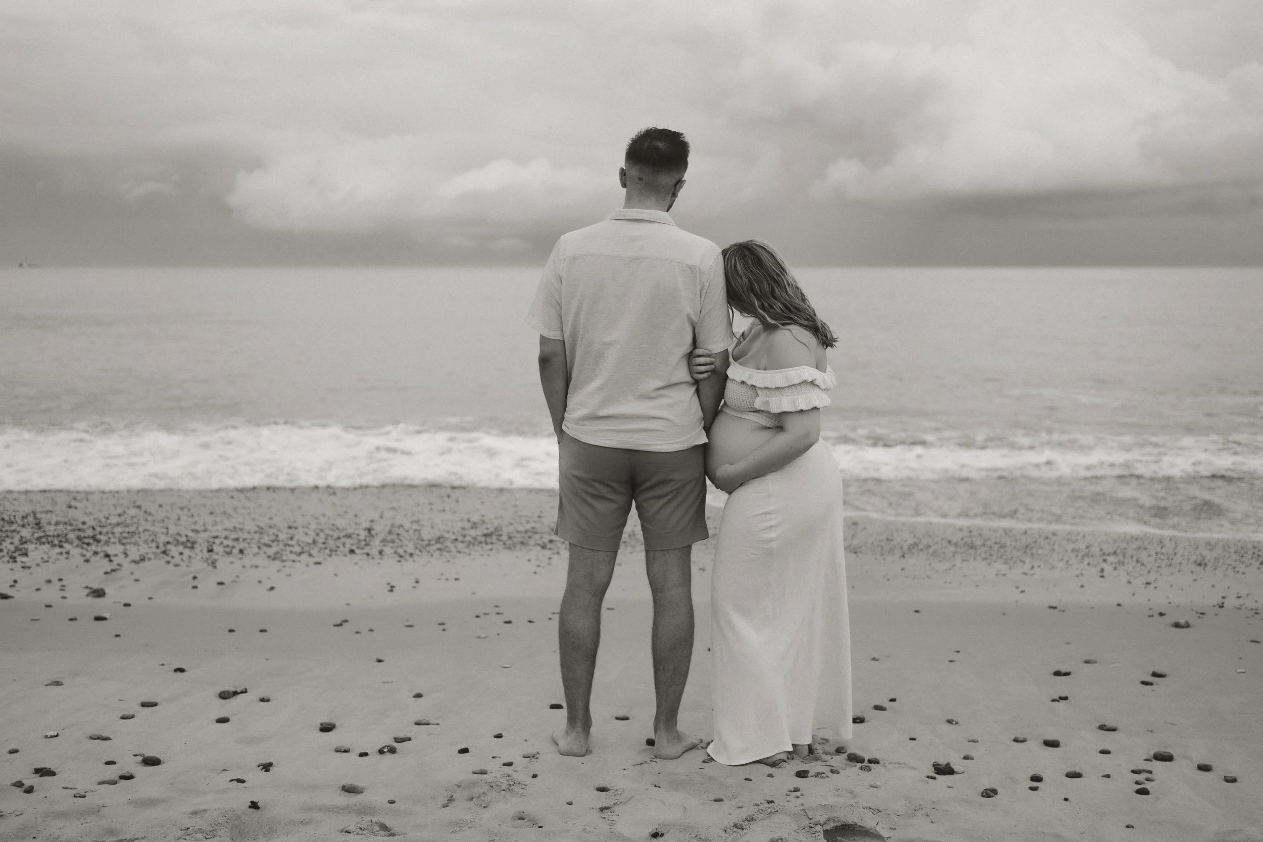 A couple stands on the beach facing the ocean, with the woman visibly pregnant, during cloudy weather.