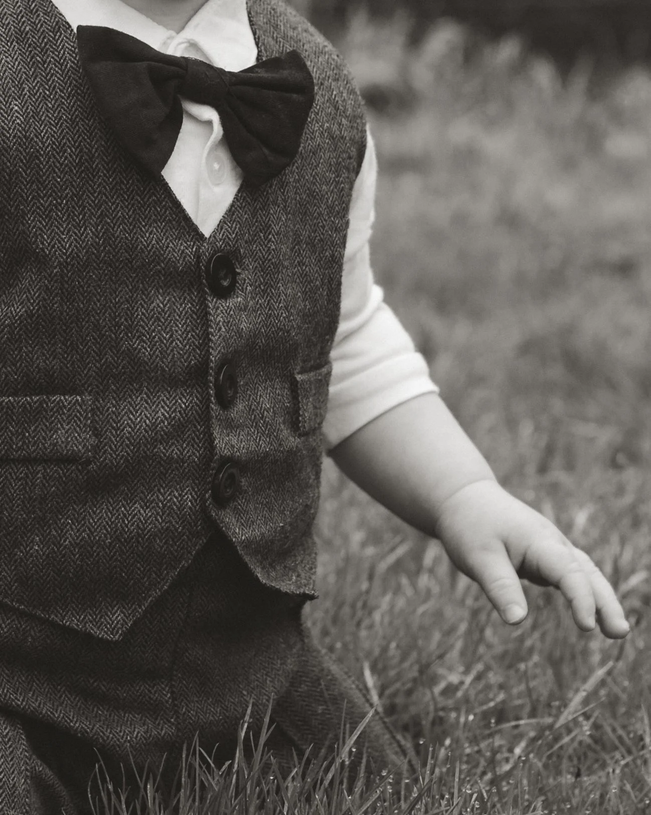 Close-up of a child's arm and hand, wearing a formal shirt, vest, and bow tie, with the child sitting on grass.