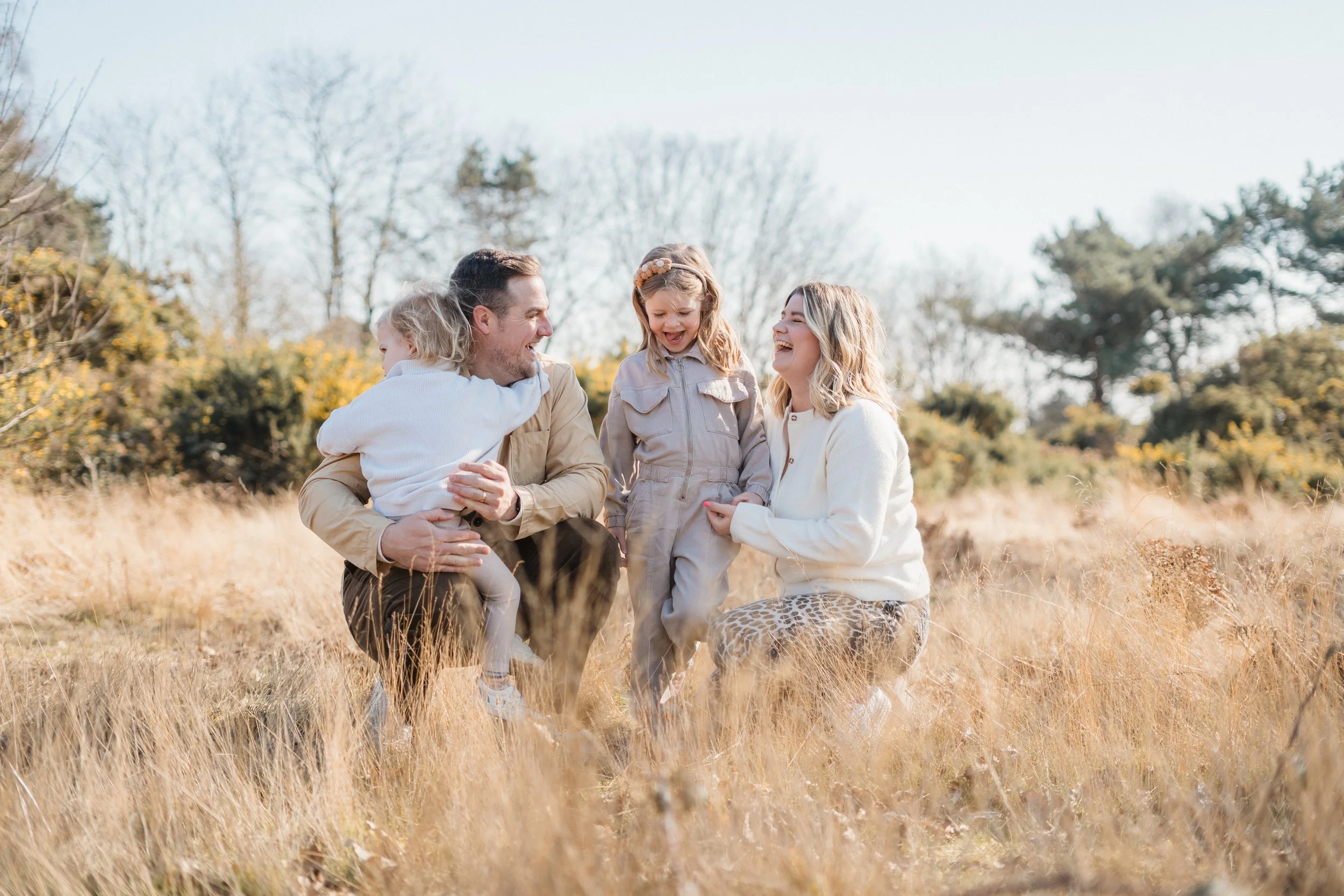 A family of four, including a father, mother, and two young girls, enjoying an outdoor moment in a grassy field during daytime, all smiling and laughing.