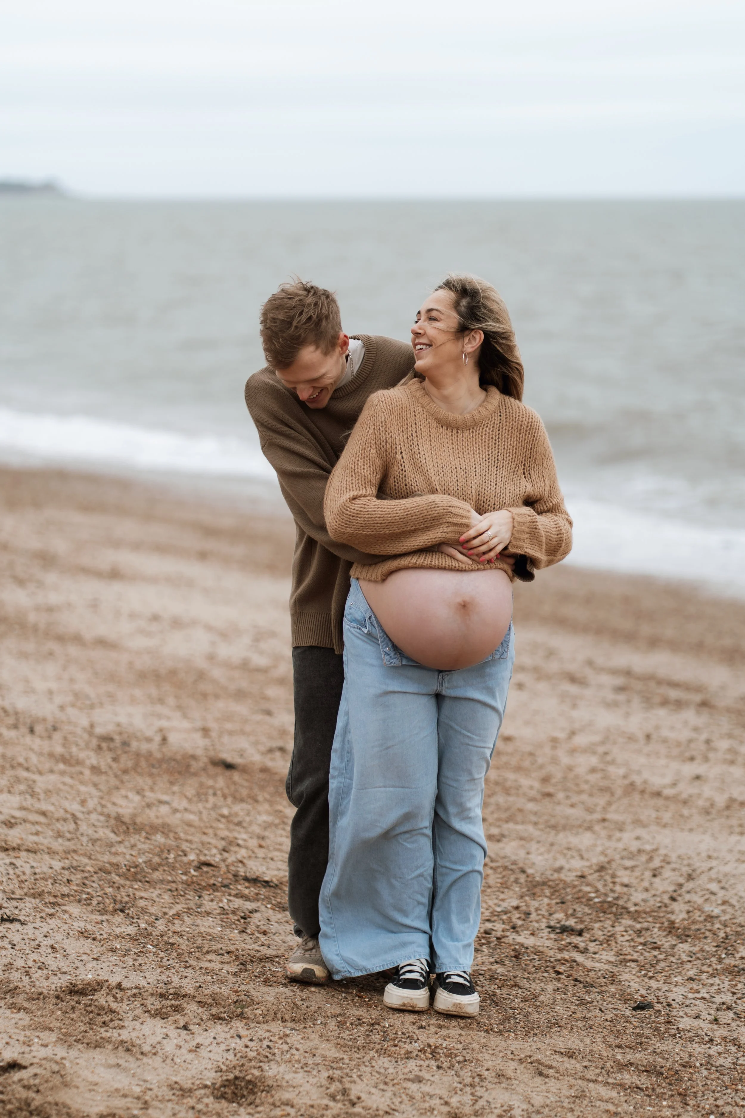A pregnant woman and a man on a beach, with the woman showing her baby bump, both smiling and enjoying a moment together.