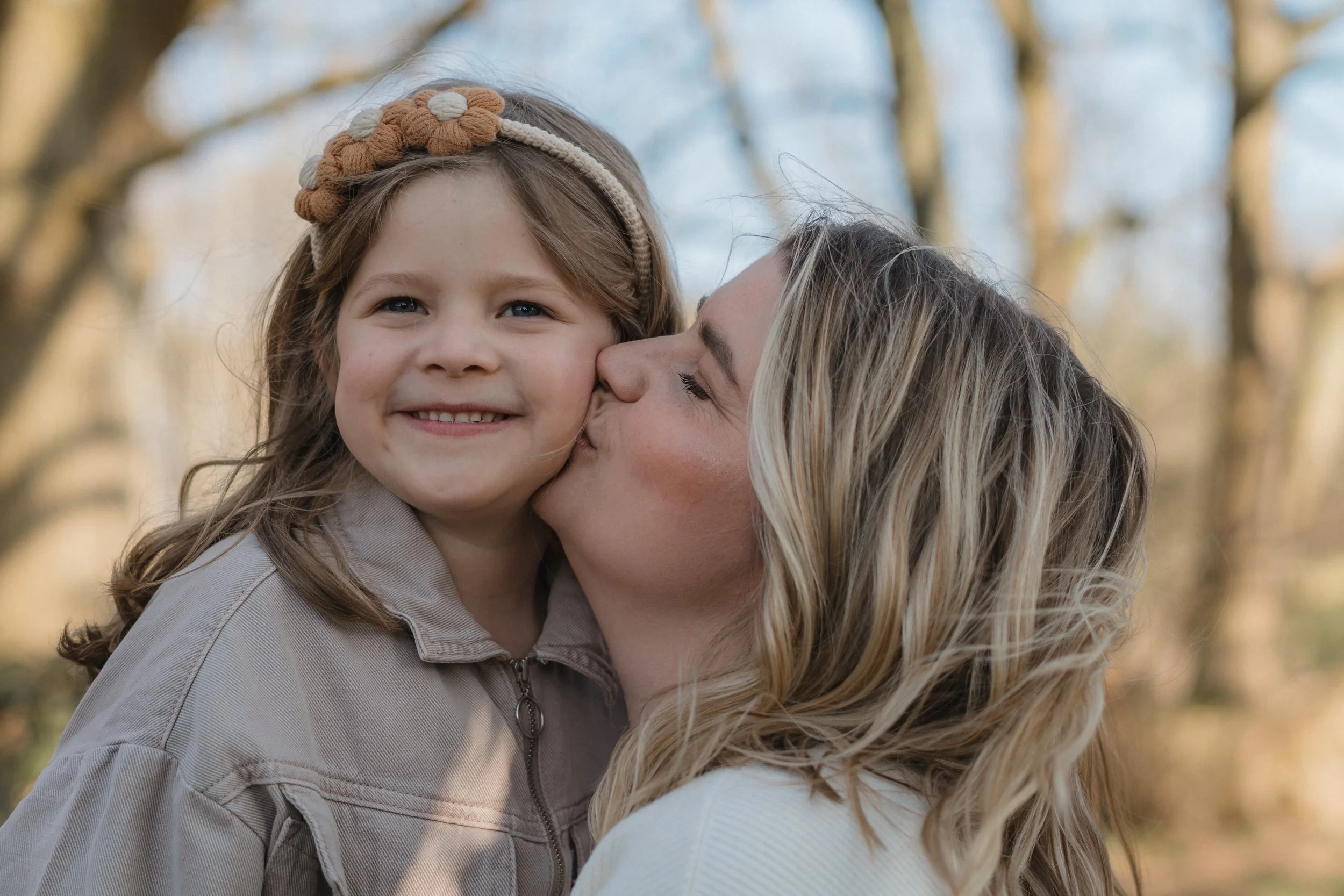 A woman with blonde hair is kissing a young girl on the cheek outdoors with trees in the background. The girl has brown hair with a flower headband and is smiling.