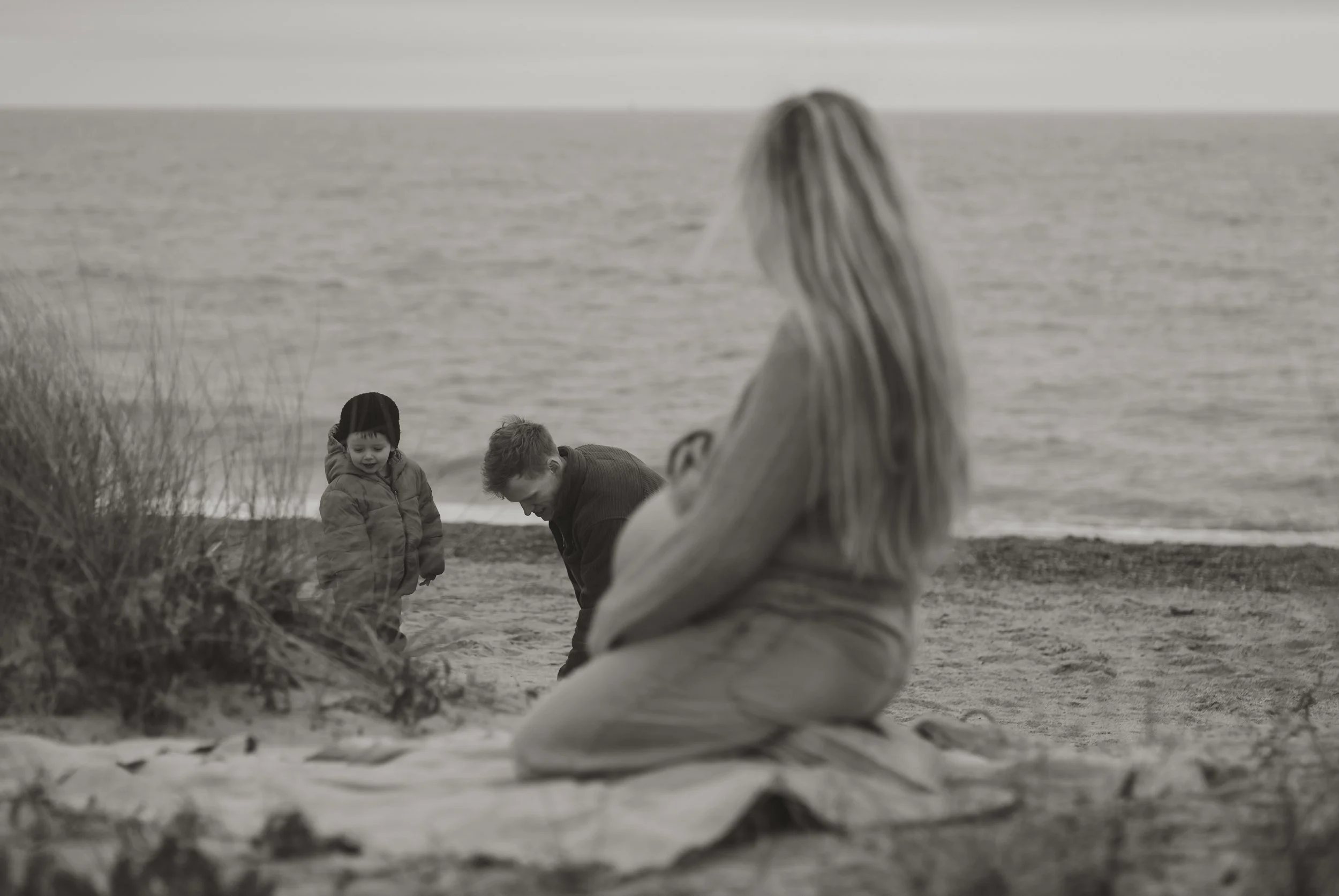 A woman with long hair kneeling on a beach, looking at two children near the shoreline, with the ocean in the background. The photo is in black and white.