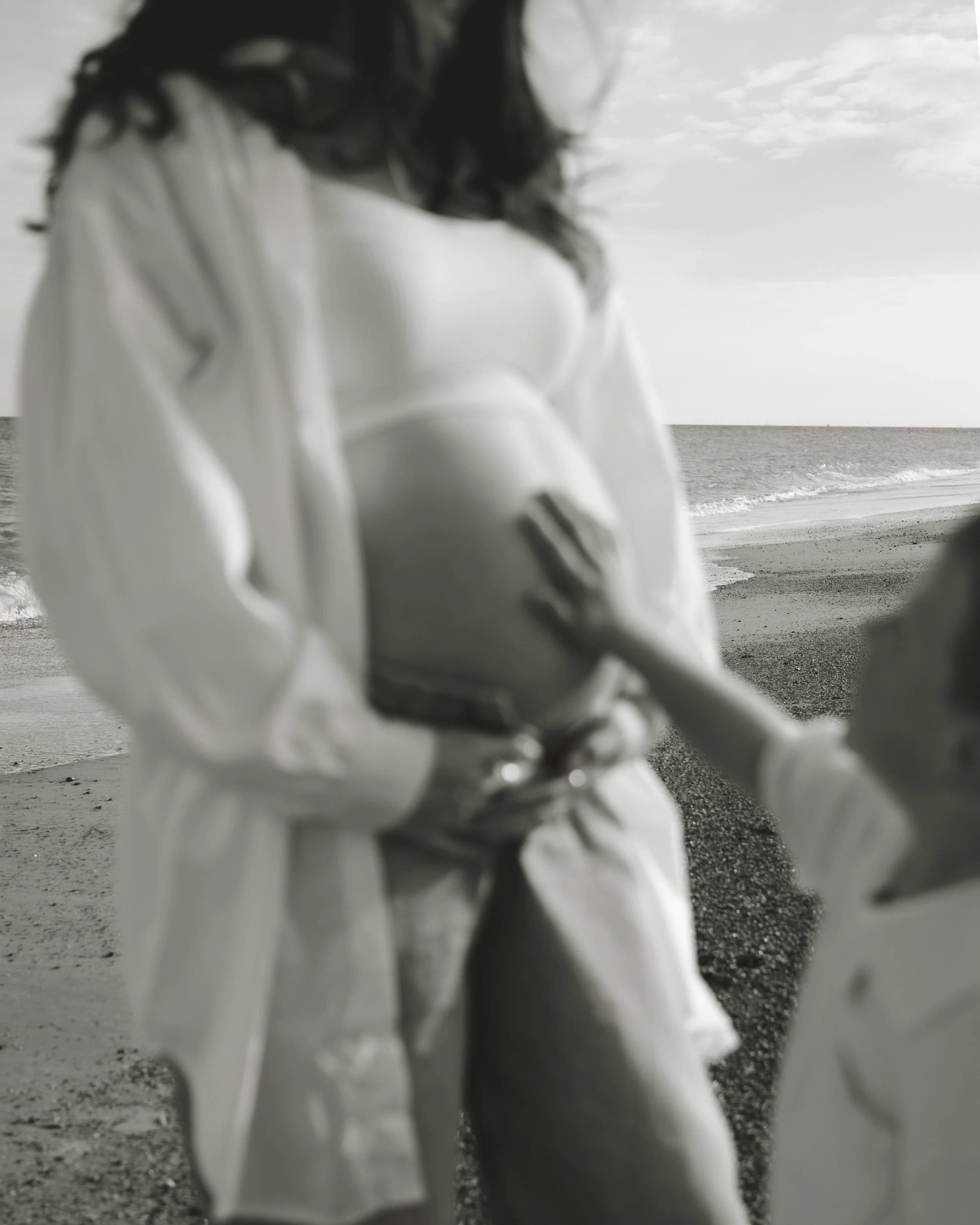 A pregnant woman standing on a beach with a hand on her belly, and a child touching her belly, with ocean and sky in the background.
