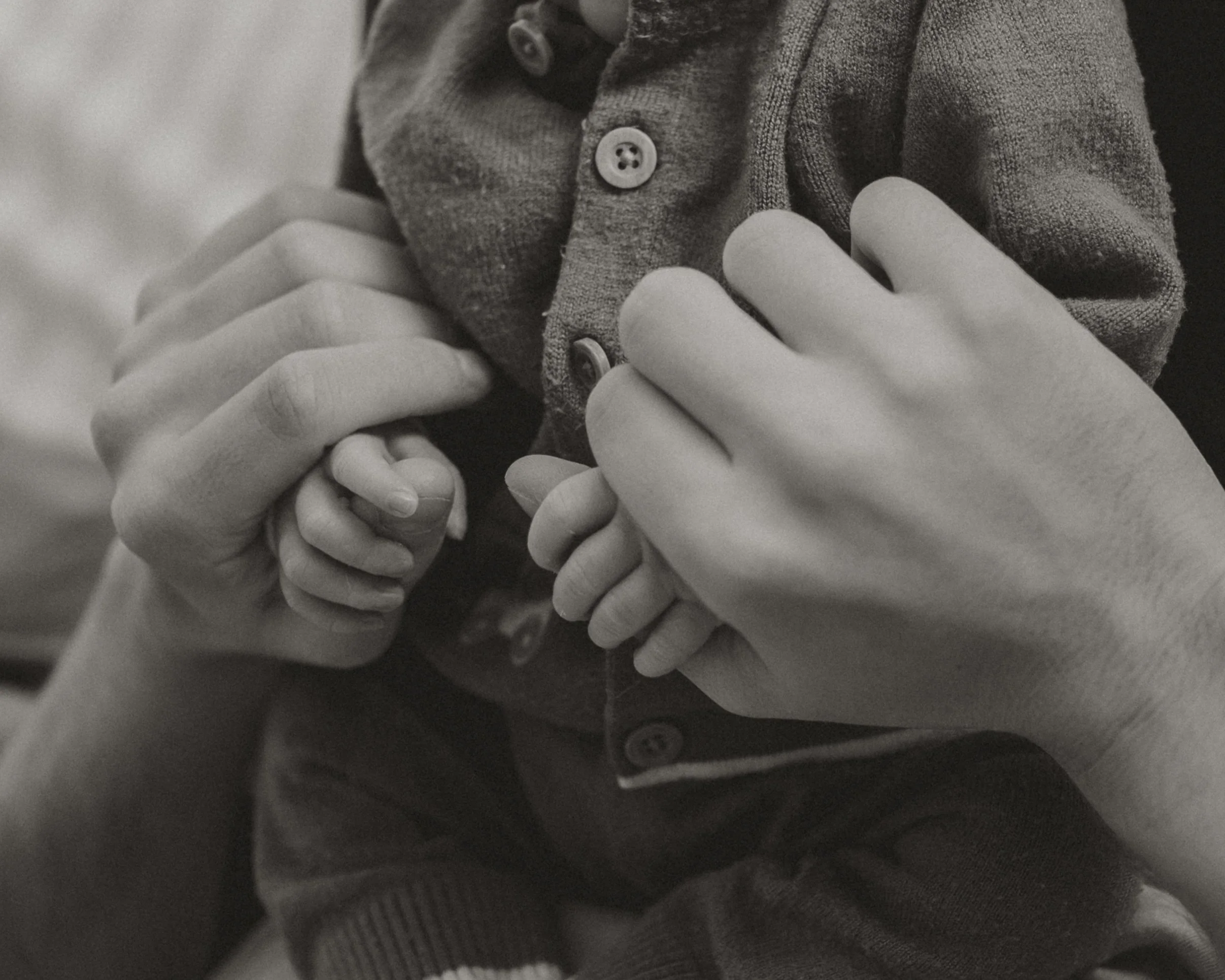 A close-up of two adult hands gently holding the small hands of a child, with the child wearing a button-up shirt.