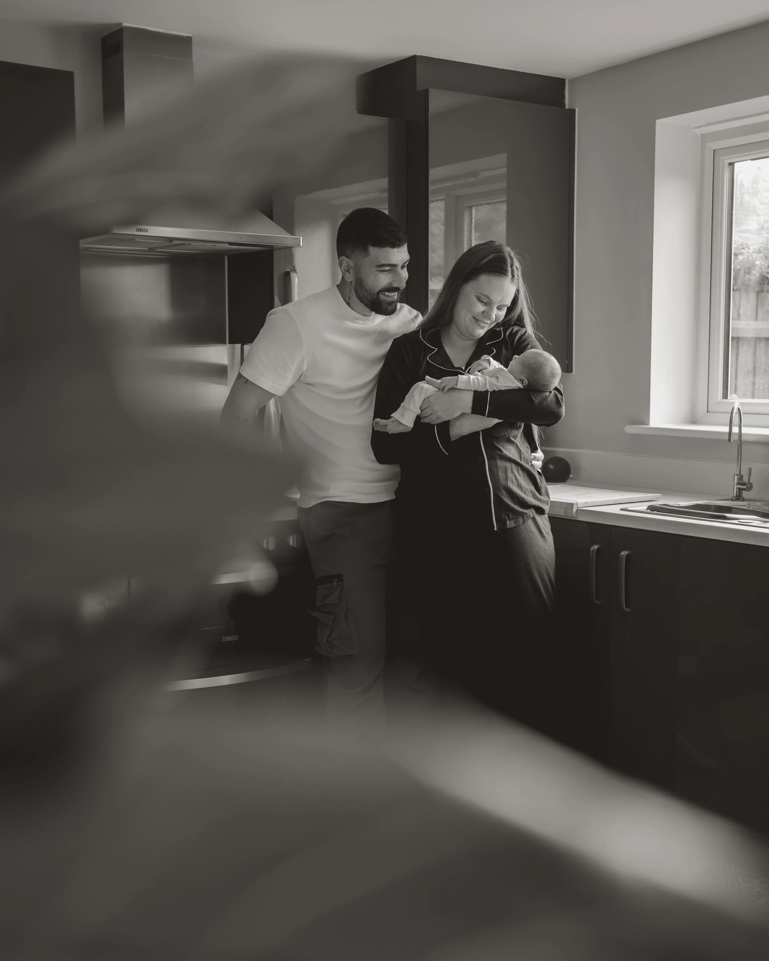 A couple stands in a kitchen, smiling as they hold a newborn baby. The scene is warm and intimate, with a window providing natural light.
