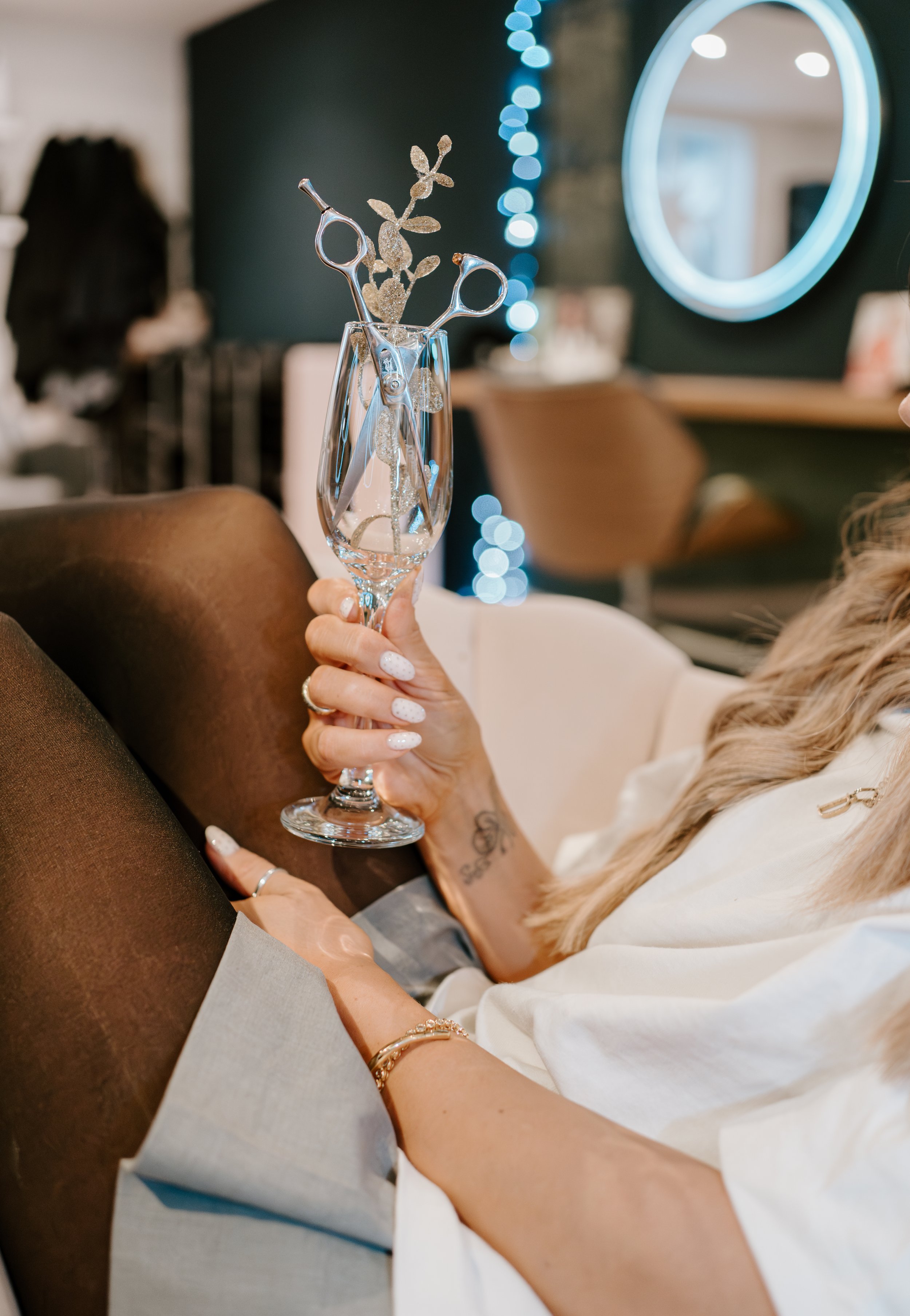 A woman with blonde hair, tattoos, and jewelry sitting on a brown chair holding a champagne flute decorated with scissors and glittery leaves, with colorful bokeh lights and a mirror in the background.