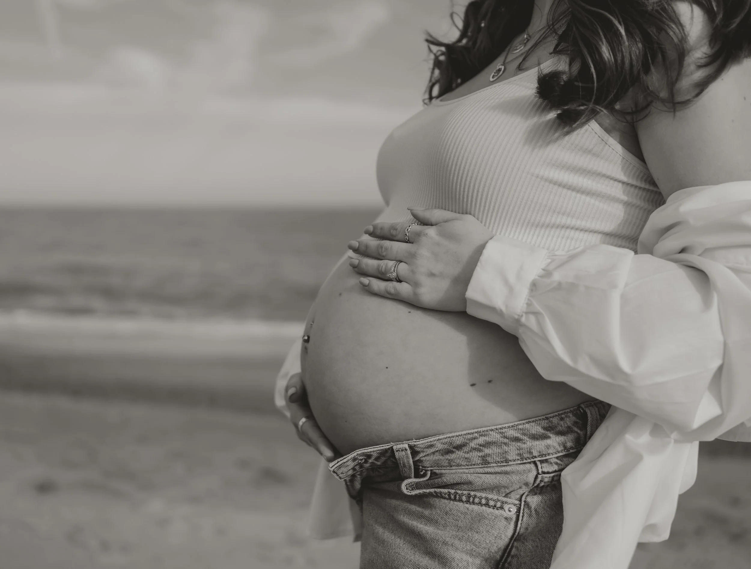 A pregnant woman stands on a beach, cradling her belly with one hand while the other rests on her stomach, showing rings on her fingers. She wears a tank top and jeans, with a white jacket draped over her shoulder. The photo is in black and white, wi