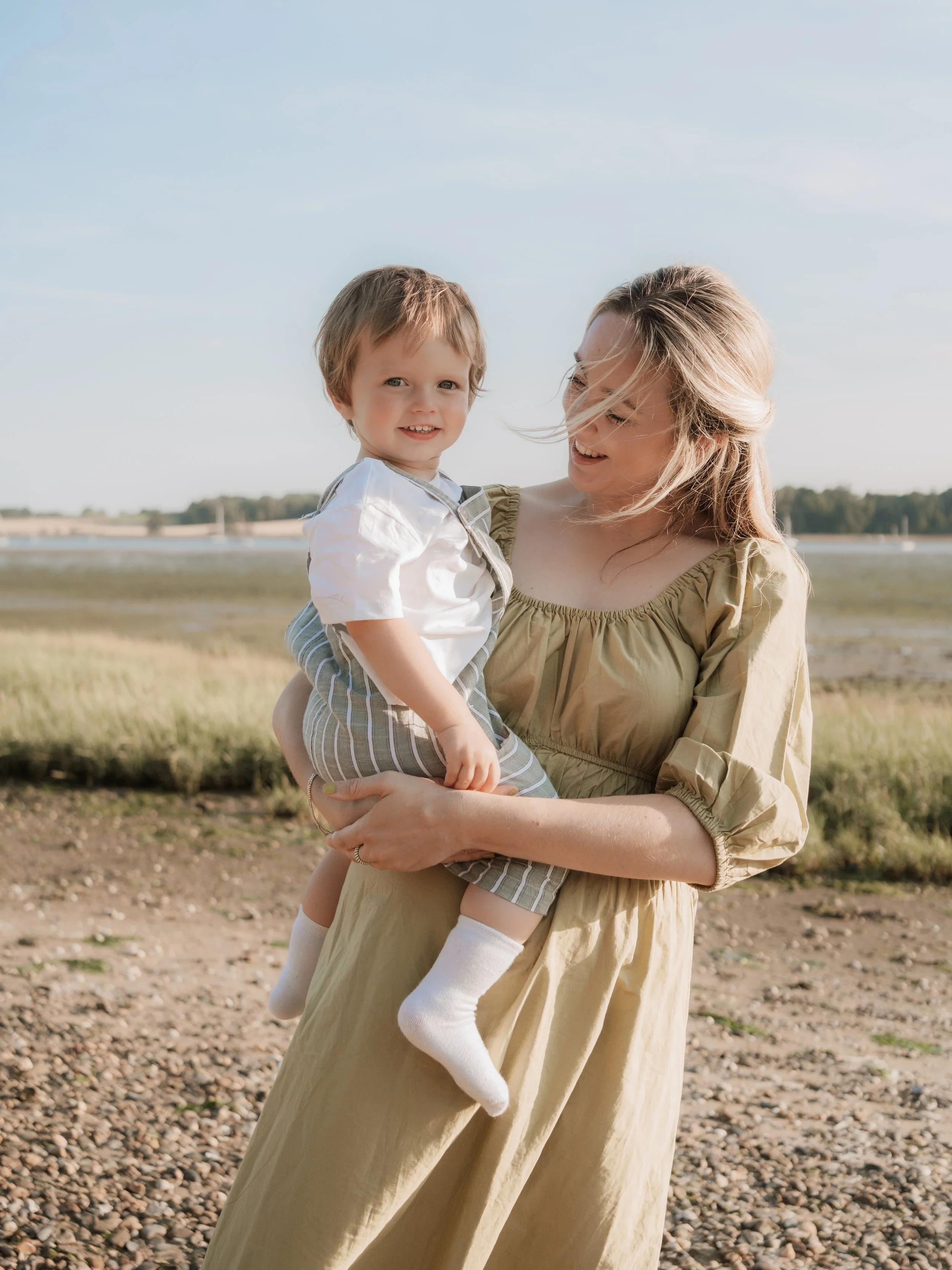 A smiling woman holding a young child outdoors near a body of water, with grass and a clear sky in the background.