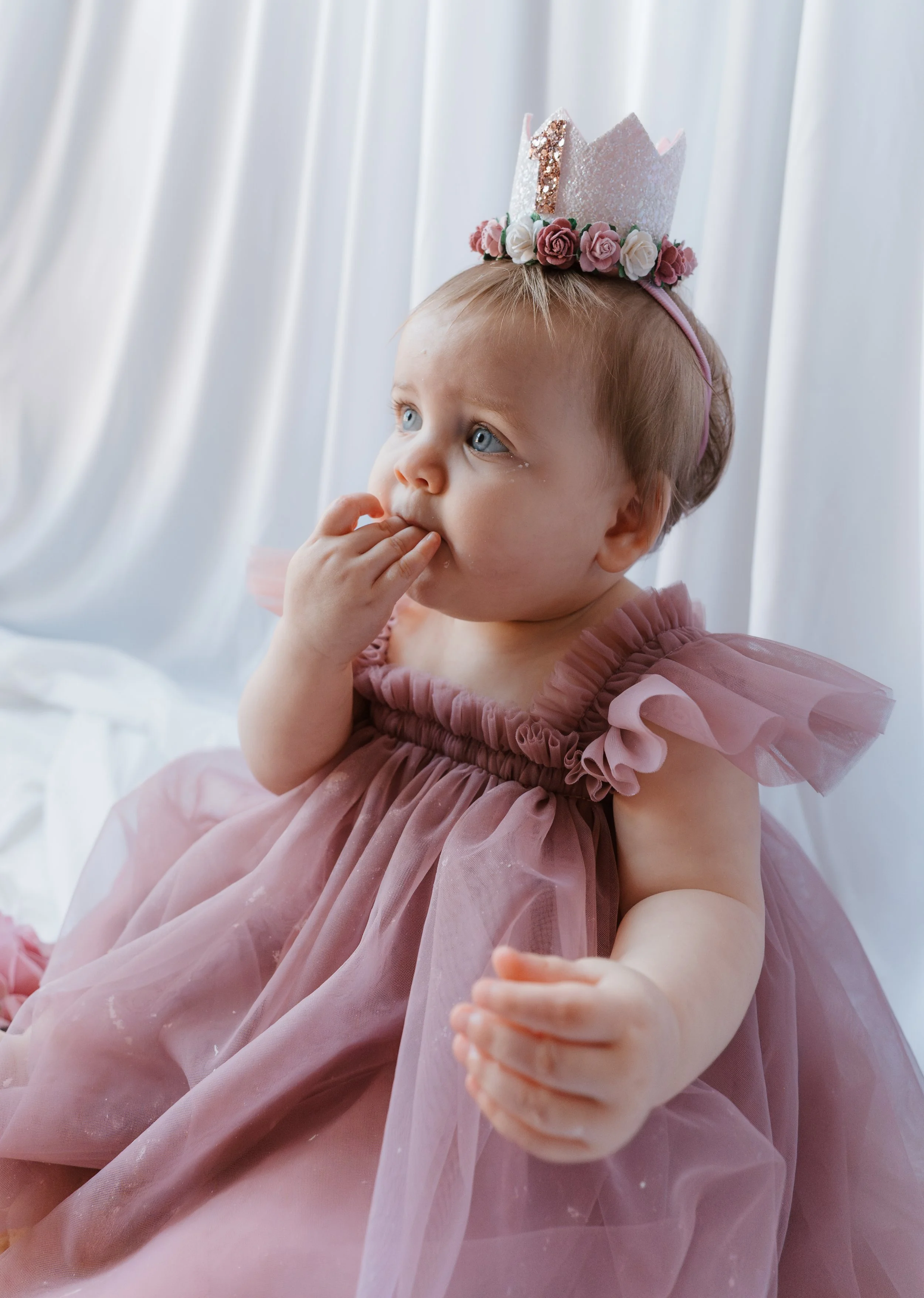 A young girl with blonde hair and blue eyes is wearing a pink dress with ruffled sleeves and a matching headband with a glittery pink crown with the number 1, and a flower border. She is sitting against a white curtain and has her fingers in her mout