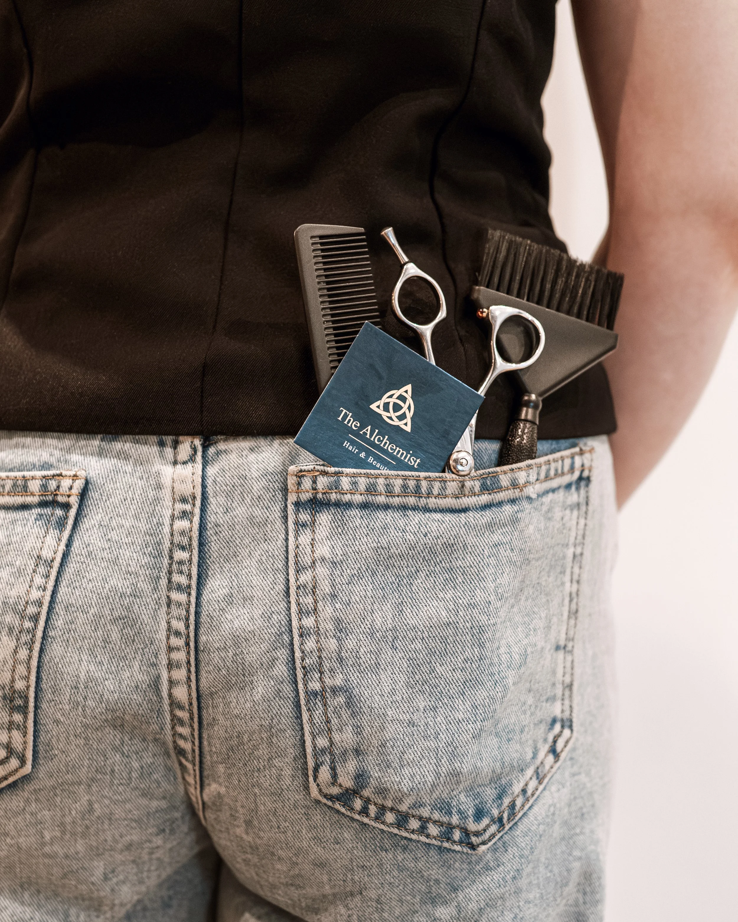 A person wearing light denim jeans with a black shirt. Hair cutting tools, including scissors, a comb, and an edge razor, are visible in the back pocket, along with a business card for The Alchemist Hair & Beauty.