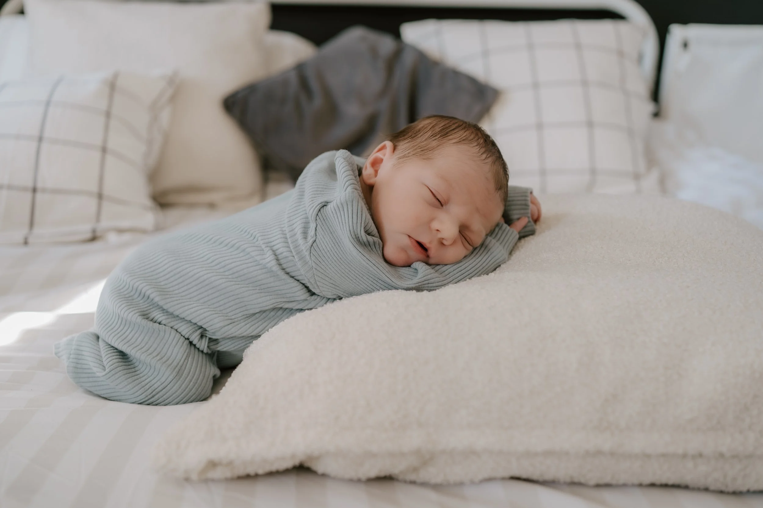 A sleeping baby lying on a cream-colored blanket on a bed with pillows in the background.