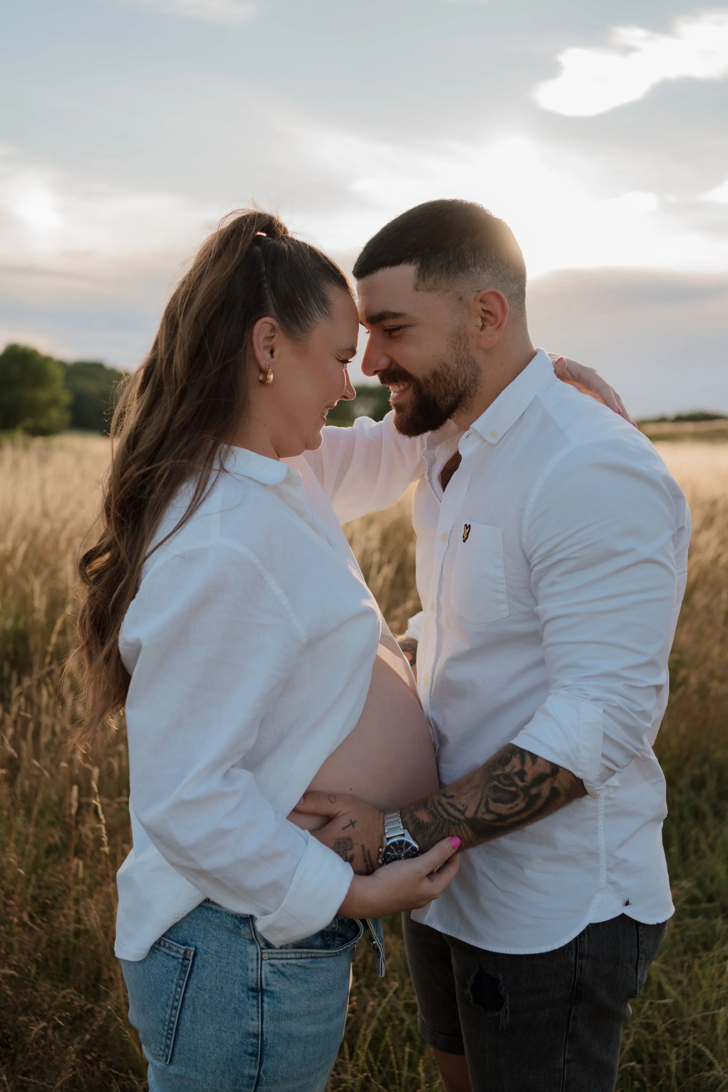 A happy couple embraces in a field during sunset, with the man touching a pregnant woman's belly and both smiling with foreheads touching.