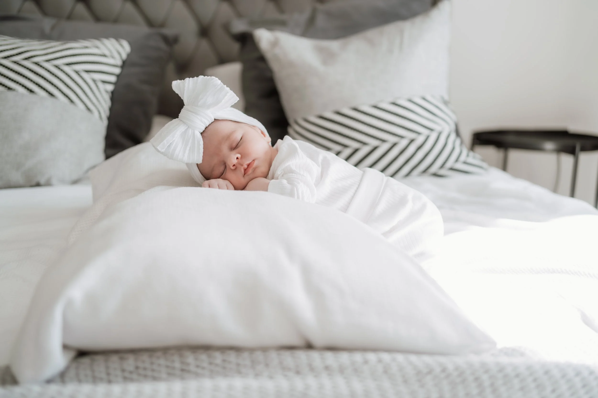 A sleeping baby with a white headband featuring a large bow, lying on a bed with white bedding and surrounded by black and white striped and solid pillows.