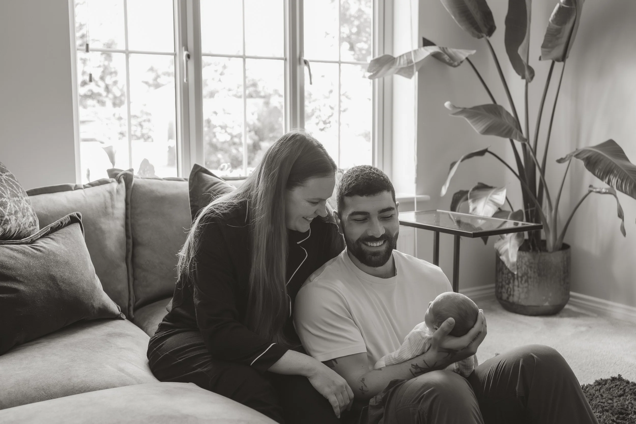 A couple sitting on a couch, looking at a newborn baby, in a bright living room with large windows, pillows on the couch, a potted plant, and a side table.