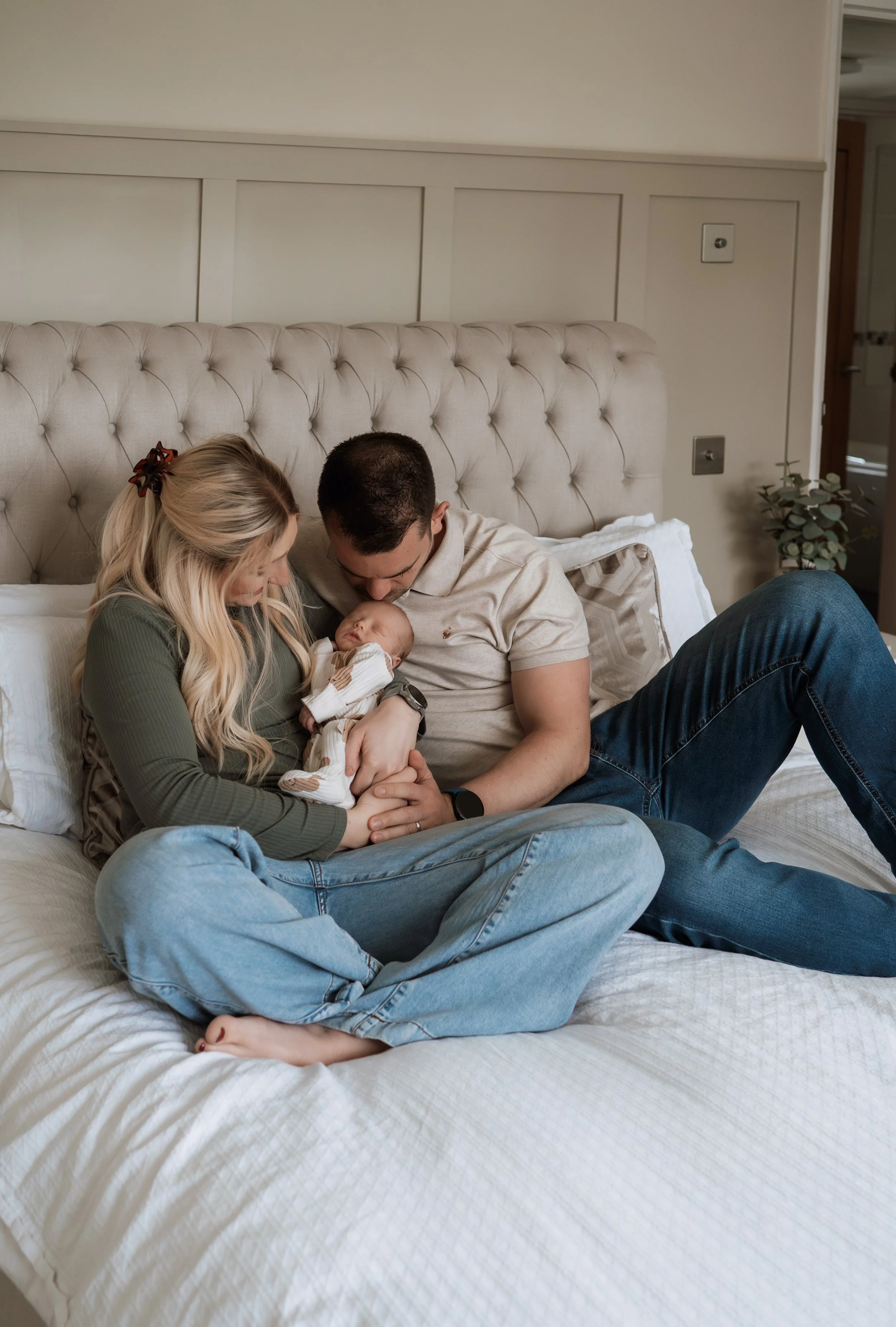 A couple sitting on a bed holding their newborn baby, gazing at the infant lovingly in a cozy bedroom.
