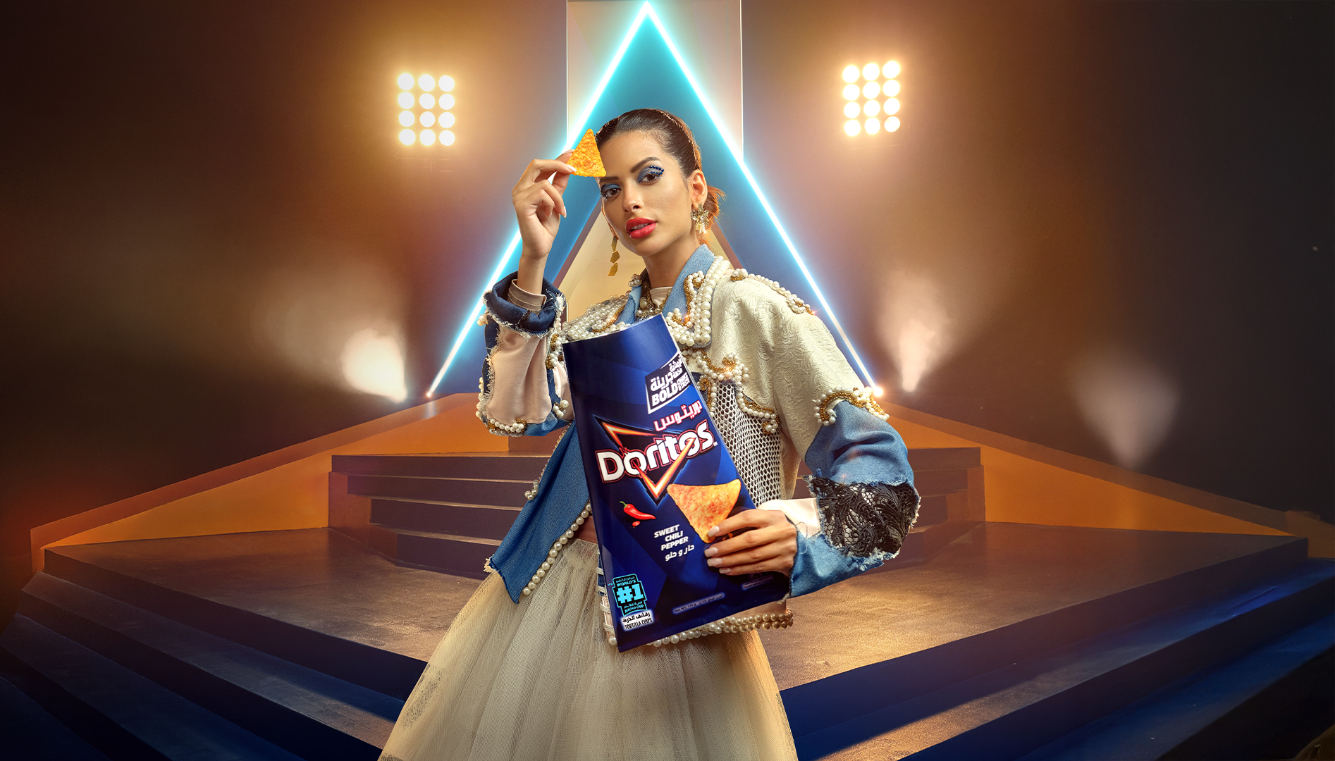 A woman dressed in an ornate, pearl-adorned blue and cream outfit is holding a bag of Doritos chips, standing in front of a stage with neon lighting and stage lights.
