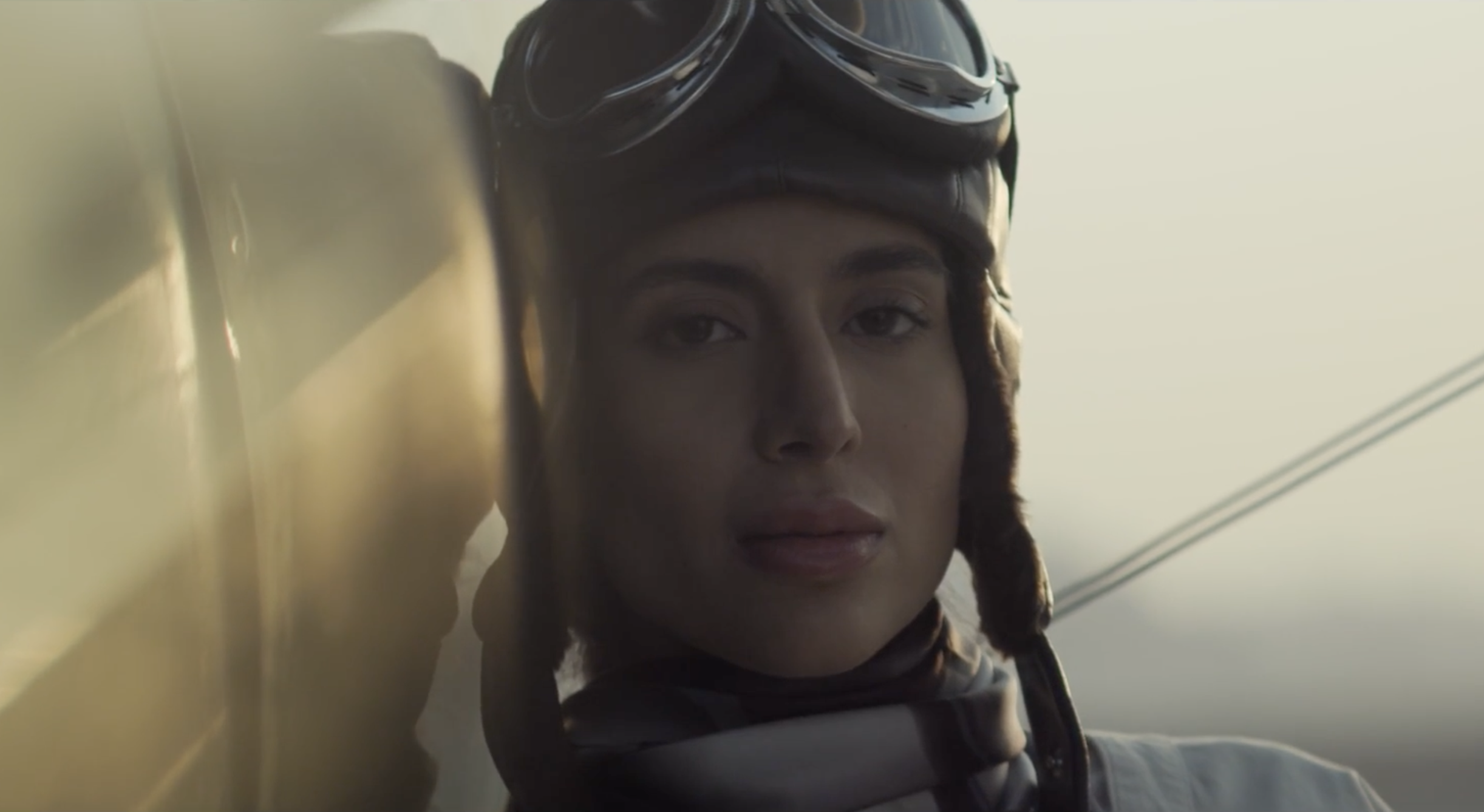 Close-up of a young woman wearing a vintage leather aviator helmet and goggles, looking thoughtfully at the camera with an outdoor background.