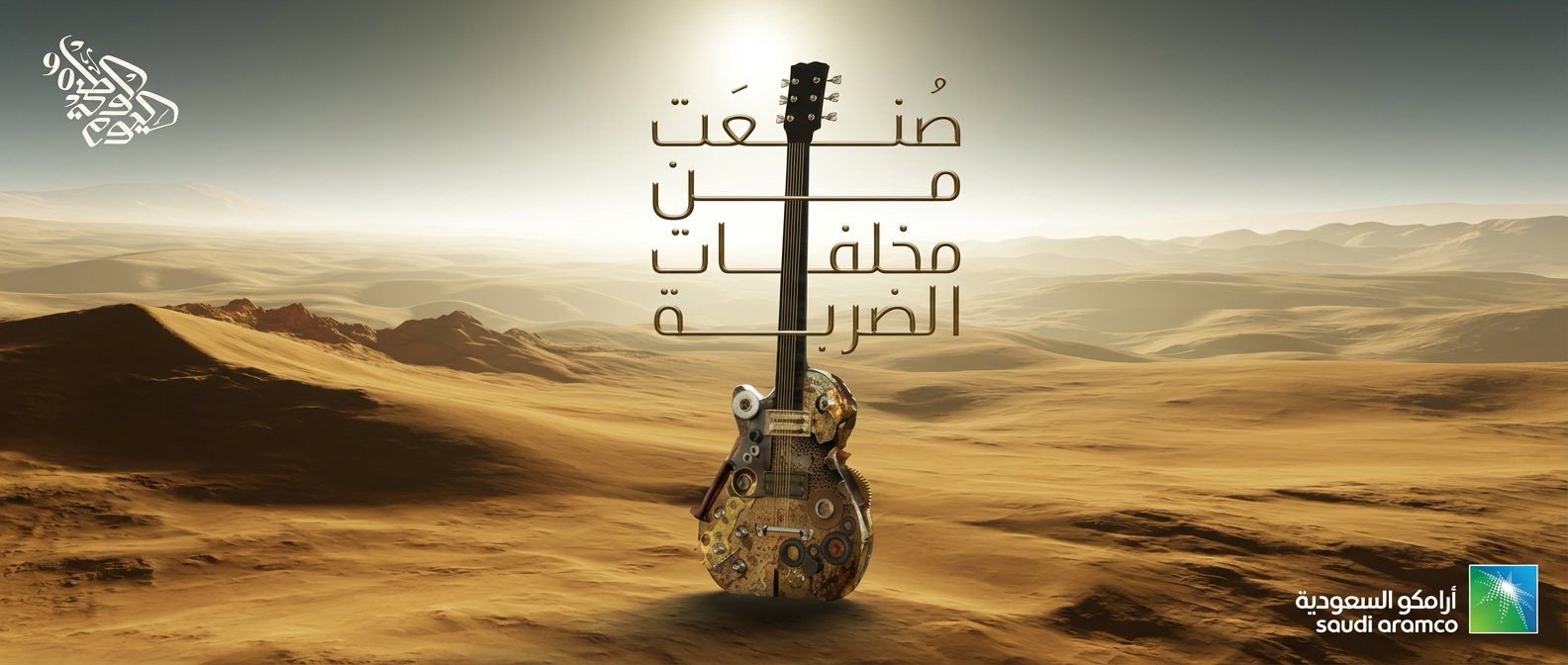 A desert landscape with mountains in the distance and a cloudy sky. A guitar with mechanical parts is standing vertically in the center of the image. Arabic text is overlaid on the guitar, and the Saudi Aramco logo is in the bottom right corner.