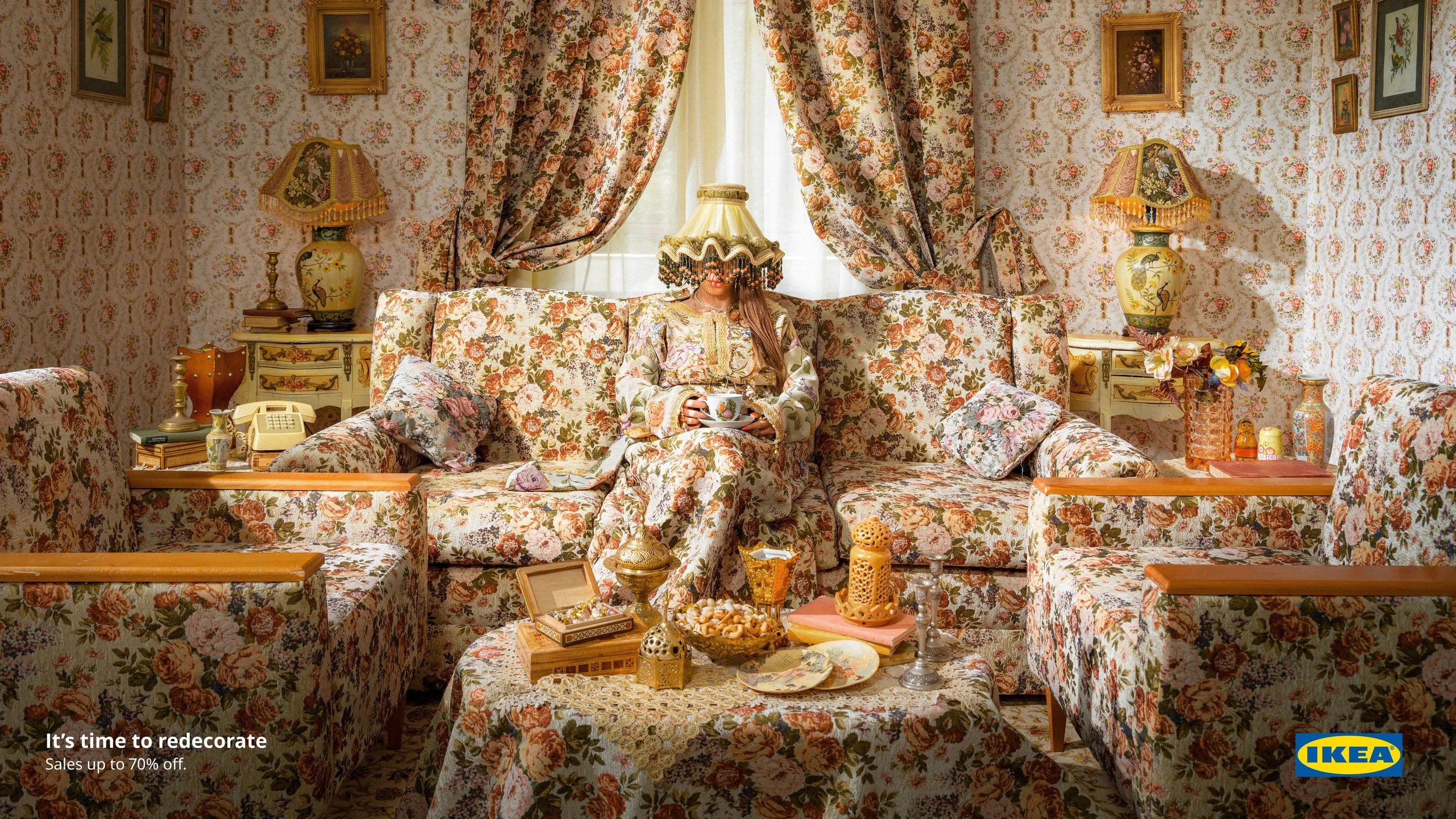 A woman sitting on a floral-patterned sofa in a room with floral wallpaper and matching curtains, holding a teacup and smiling underneath a decorative lampshade on her head, surrounded by vintage furniture and decorations.